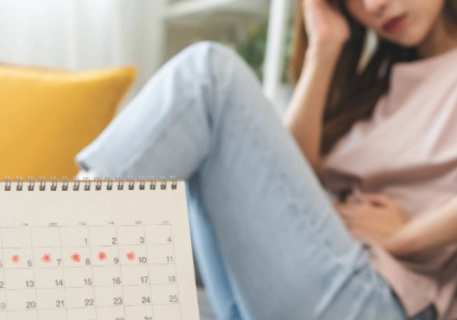A woman holding her lower abdomen with a concerned look, possibly near a calendar (to suggest menstrual tracking)