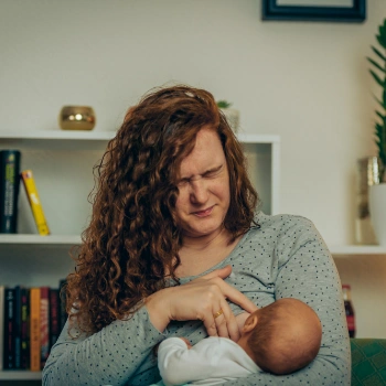 A woman breastfeeding her baby in a living room, showing signs of discomfort due to nipple pain