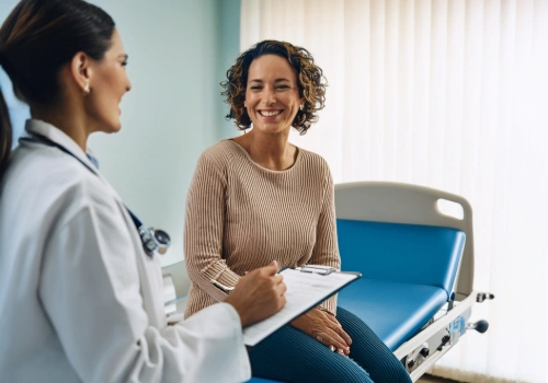 A mom in her 30s–50s smiling during a routine doctor’s visit, sitting on an exam table talking with a female provider