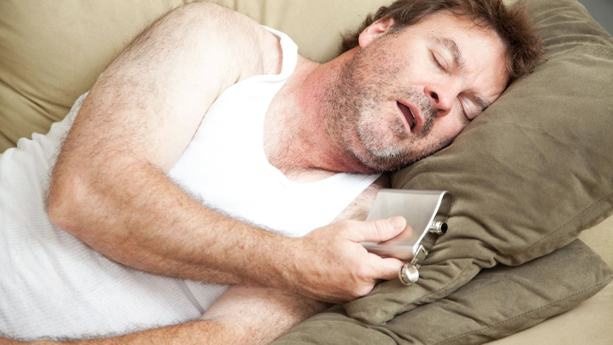 Man asleep on the couch at night with a flask of alcohol in his hand
