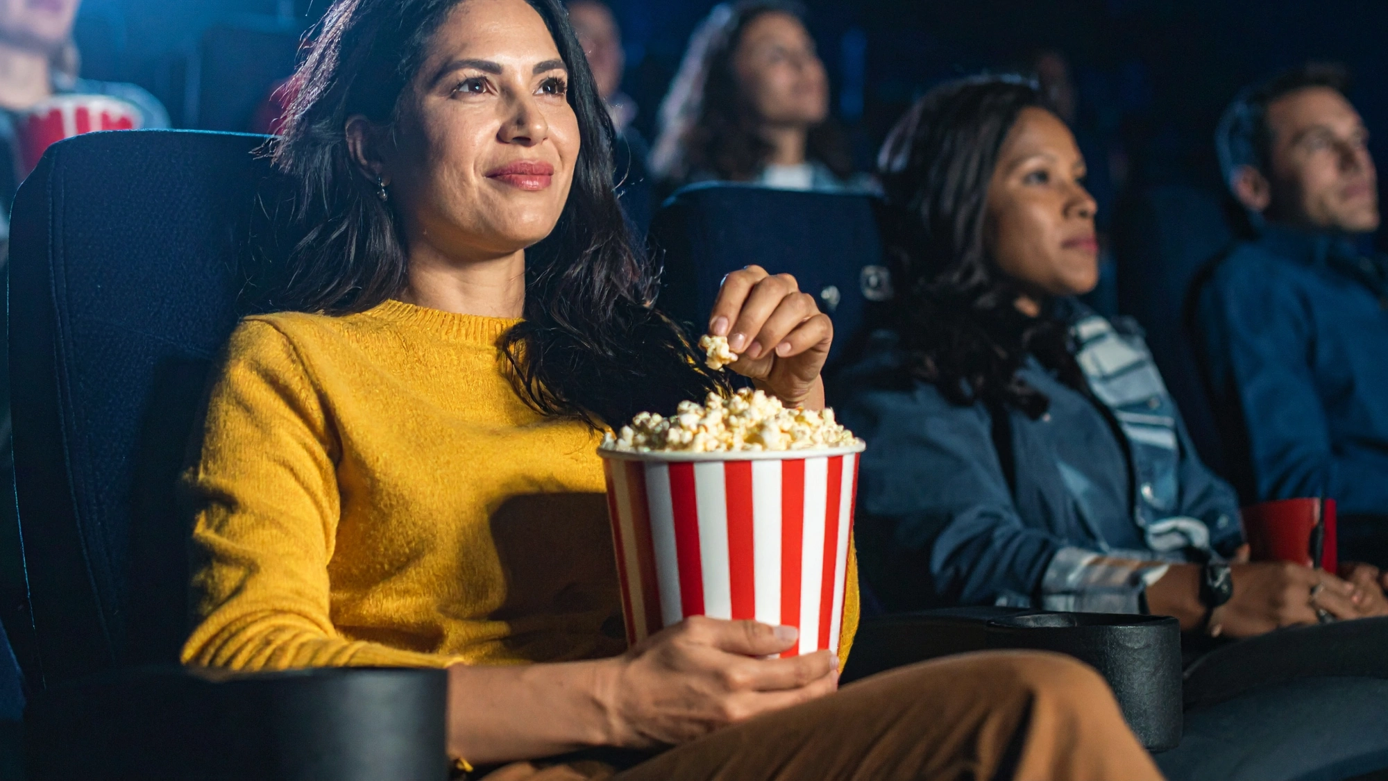 Adult woman eating salty popcorn from a classic striped popcorn bucket while seated in a dark movie theater
