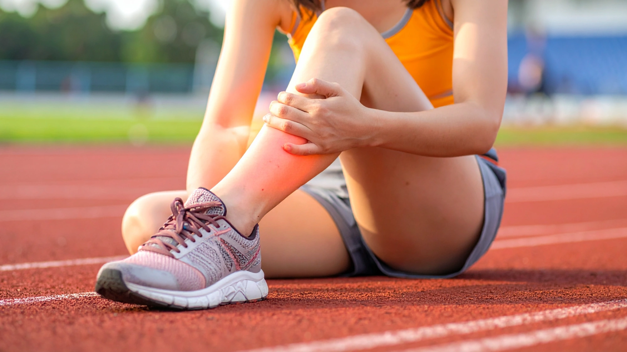 Teen girl sitting on a running track holding her lower leg due to shin splint pain.
