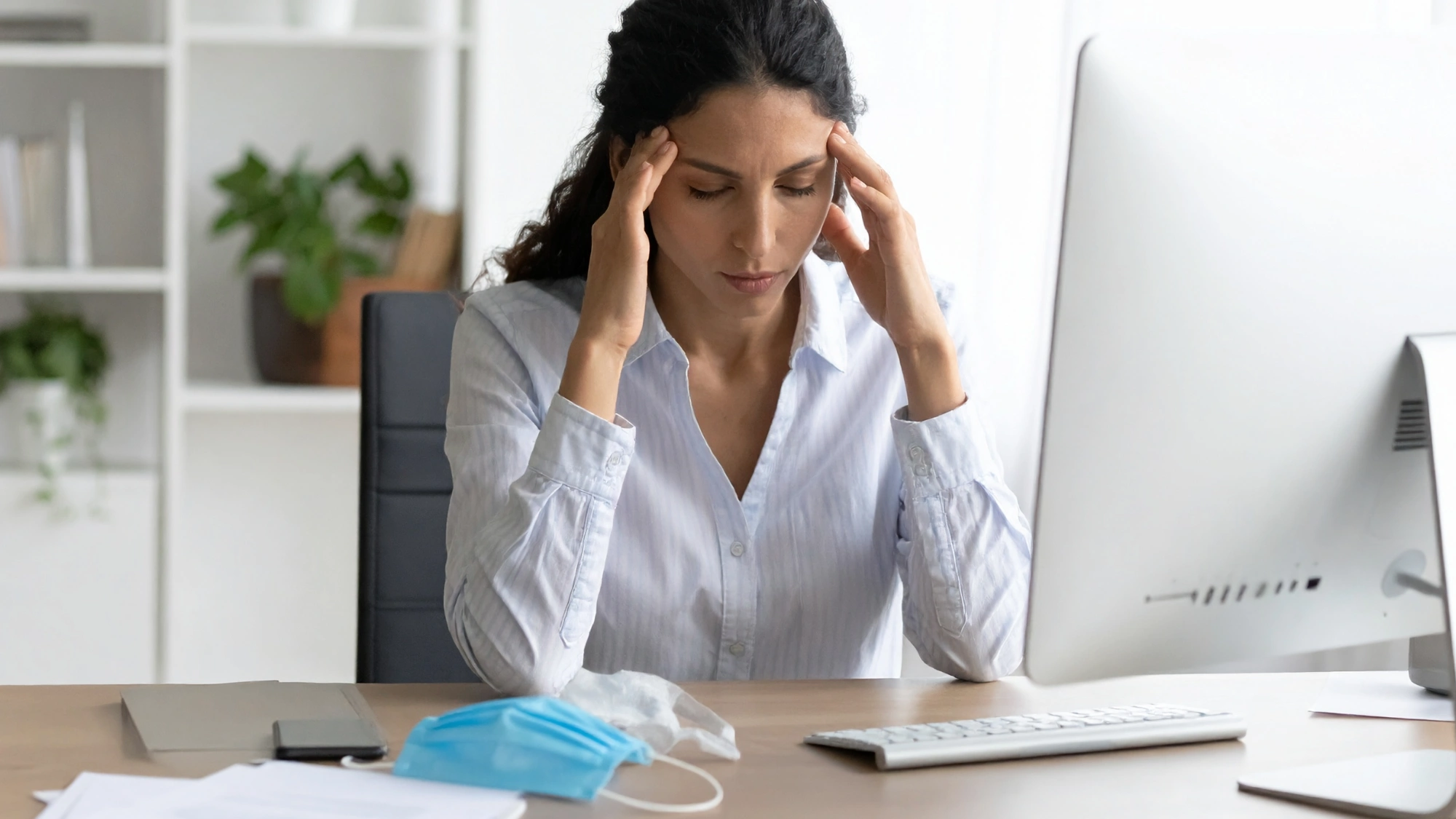 Woman sitting at her desk with her hands on her head and a face mask beside her computer while feeling sick at work.