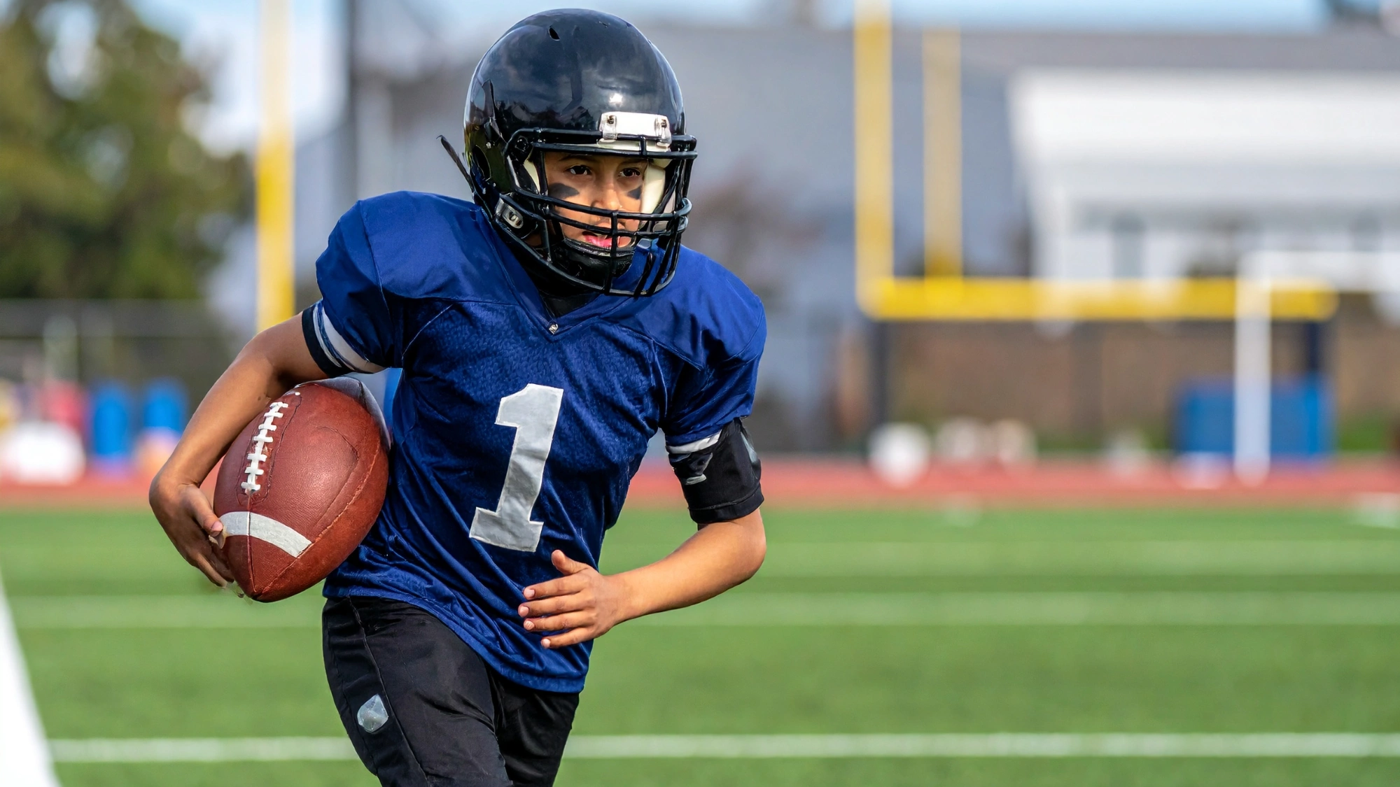 Young kid running with a football in protective gear on a multi-purpose field.