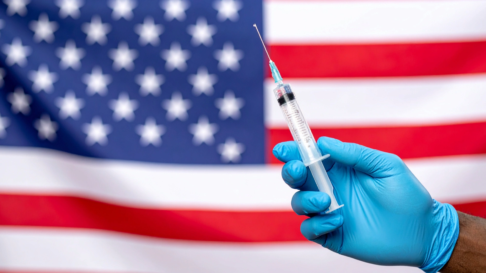 Gloved hand holding a COVID-19 booster vial in front of an American flag.