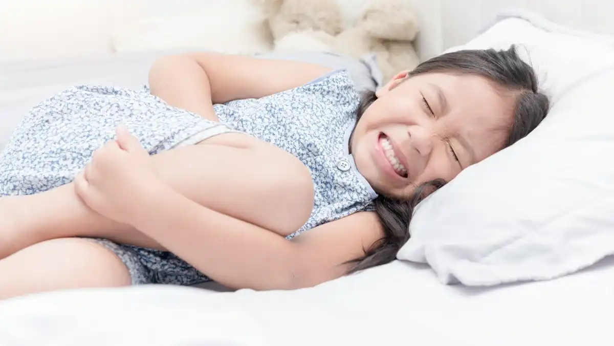 Preteen girl lies in bed clutching her stomach in discomfort, illustrating abdominal pain linked to a nervous stomach or anxiety.