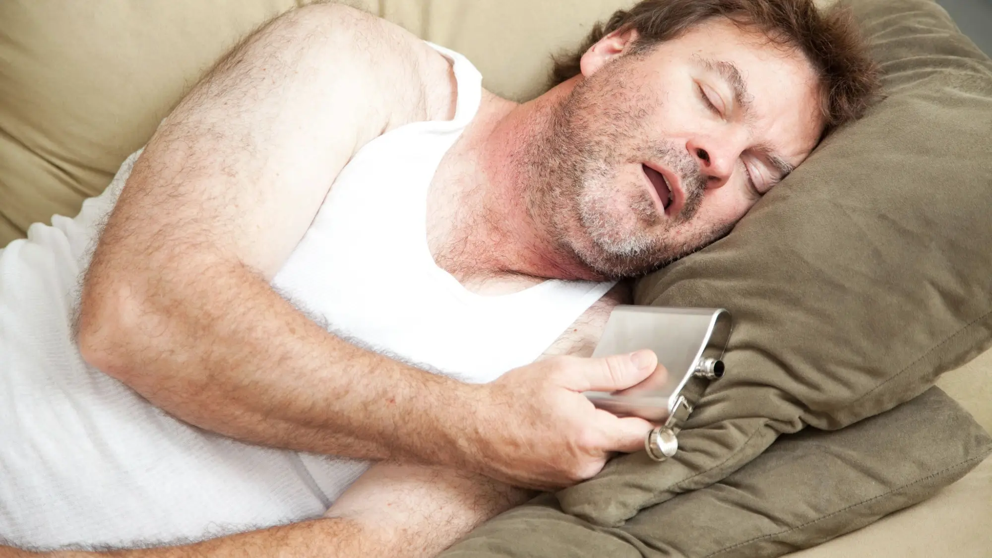 Man asleep on the couch at night with a flask of alcohol in his hand