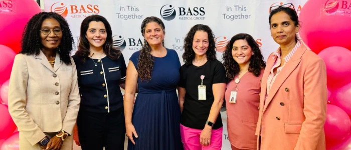 Six BASS Medical Group team and leadership members standing together at a Breast Cancer Awareness event with pink balloons and BASS Medical Group signage in the background.