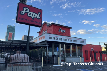 Exterior of a restaurant named Papi Tacos & Churros under a partly cloudy sky with an outdoor patio area on the left and a parking lot.