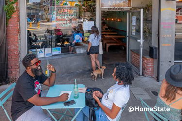 Outdoor seating area of a restaurant which features a neon window sign reading "Home of the Breakfast Taco." And two people are seated at a light blue table talking, while another person stands with a dog near the open glass entrance.