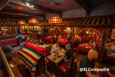 A dimly lit, vibrant indoor dining room of a restaurant features classic Mexican-style decor, including terracotta roof tiles over red leather booths, hanging stained-glass light fixtures with multiple groups of patrons are seated at tables and booths eating.
