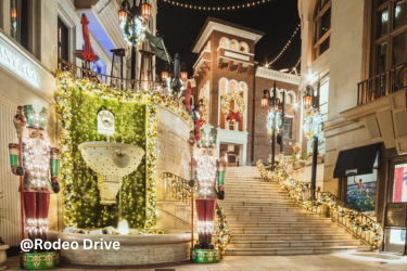 A staircase on Rodeo Drive is lined with glowing holiday garlands, decorated lampposts, and festive storefronts at night.