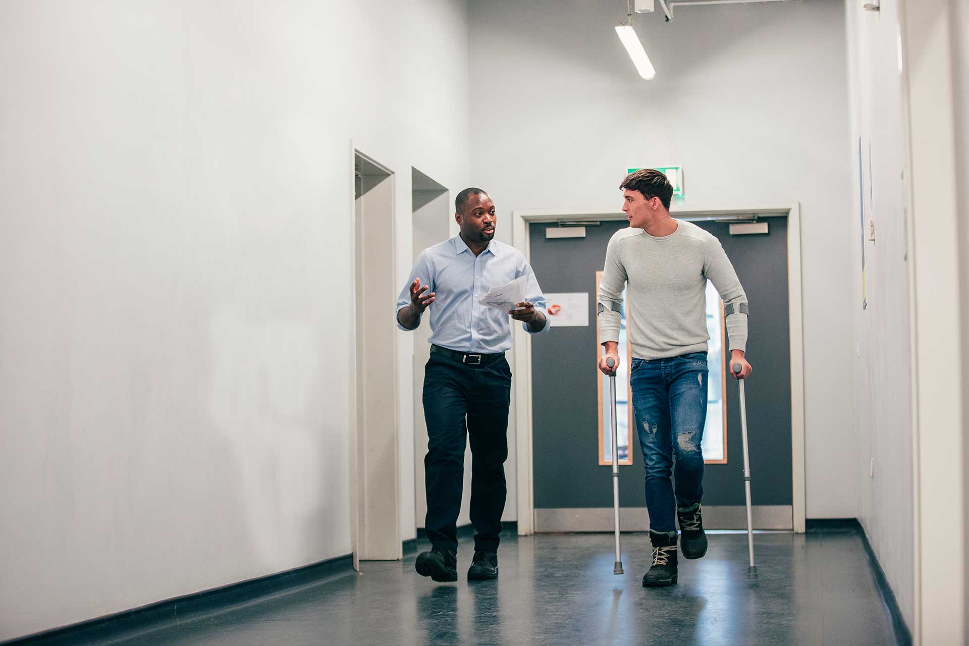 A person with crutches walking alongside another person in a hallway.