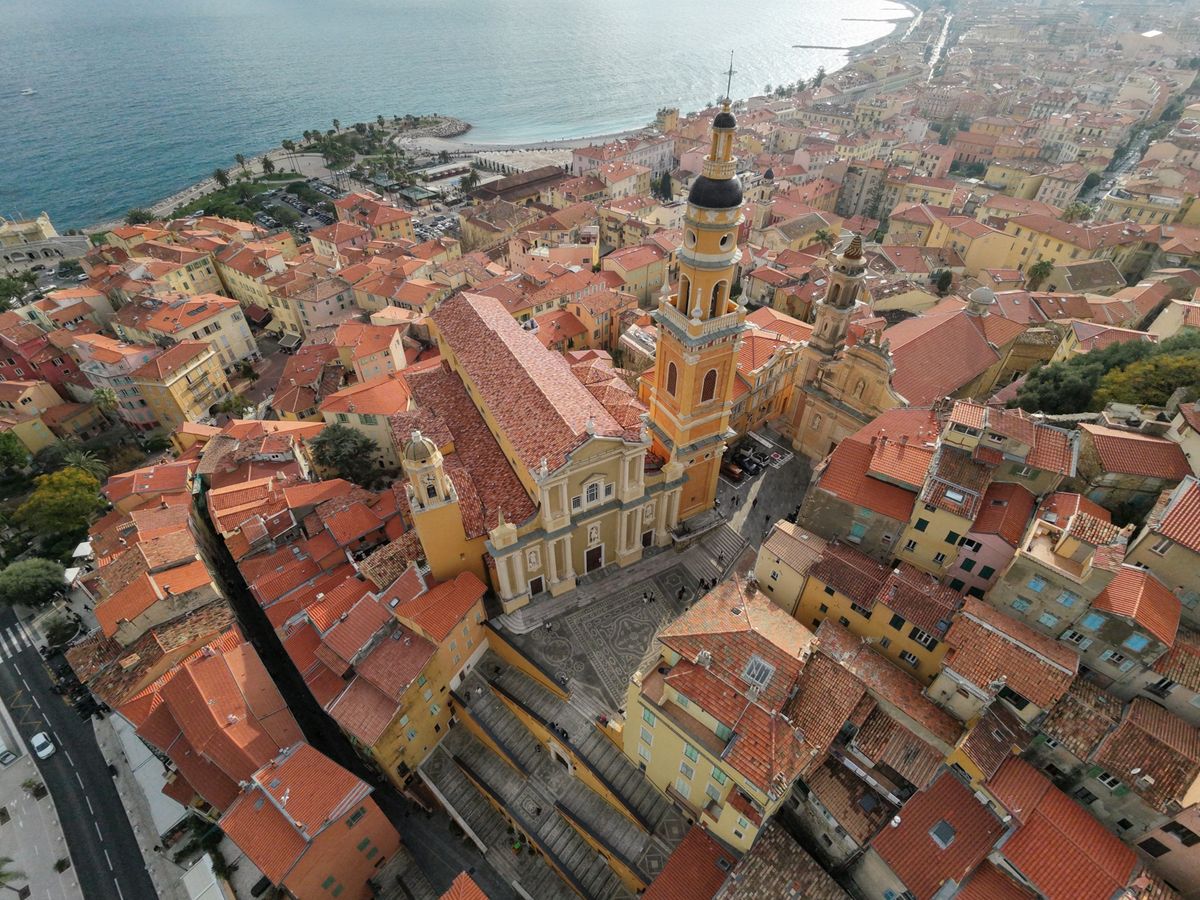 Narrow medieval alleyway in Menton old town with golden and orange facades, early morning sunlight casting long shadows, tourists walking uphill on cobblestones toward baroque church towers visible in background, colorful shutters and flower pots on windowsills, authentic Côte d'Azur architecture