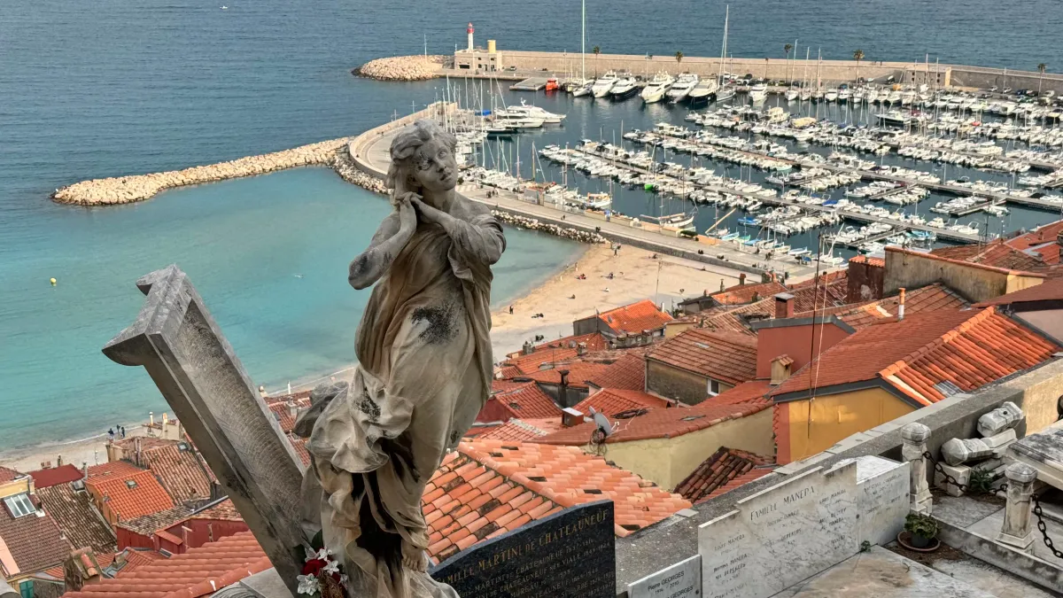 Panoramic view from Menton old cemetery overlooking the turquoise Mediterranean bay, colorful buildings cascading down the hillside, Italian border visible in the distance, alpine mountains in the background, morning light, travel photography perspective, real photographic scene