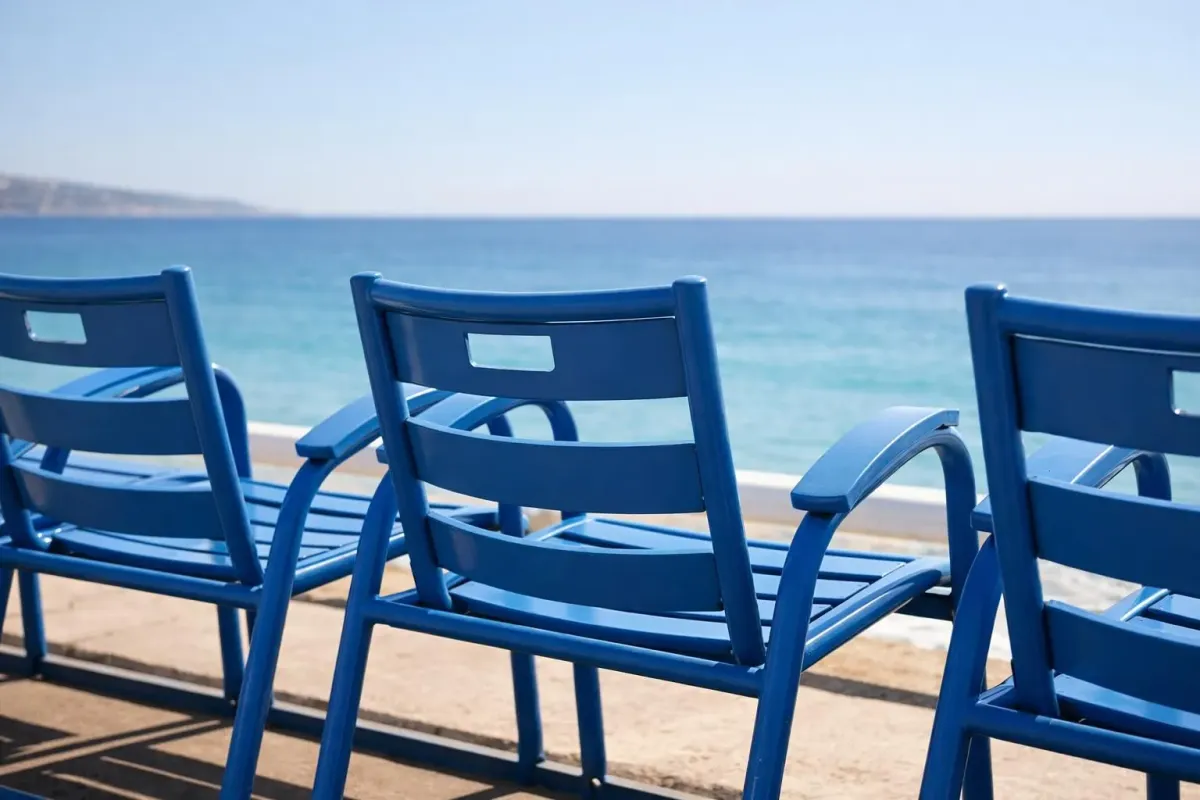 Promenade des Anglais in Nice with iconic blue chairs lined along Mediterranean coastline, palm trees swaying, tourists walking and cycling on sunny afternoon, turquoise sea in background, typical French Riviera atmosphere without any text or signs