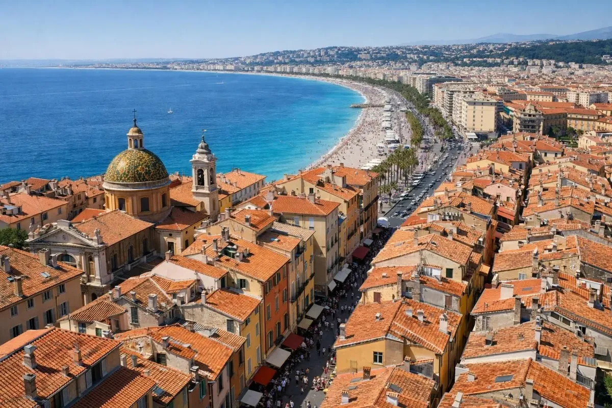 Aerial view of Nice old town with terracotta rooftops, narrow streets leading to turquoise Mediterranean sea, Promenade des Anglais in background, tourists walking along colorful building facades, bright afternoon sunlight