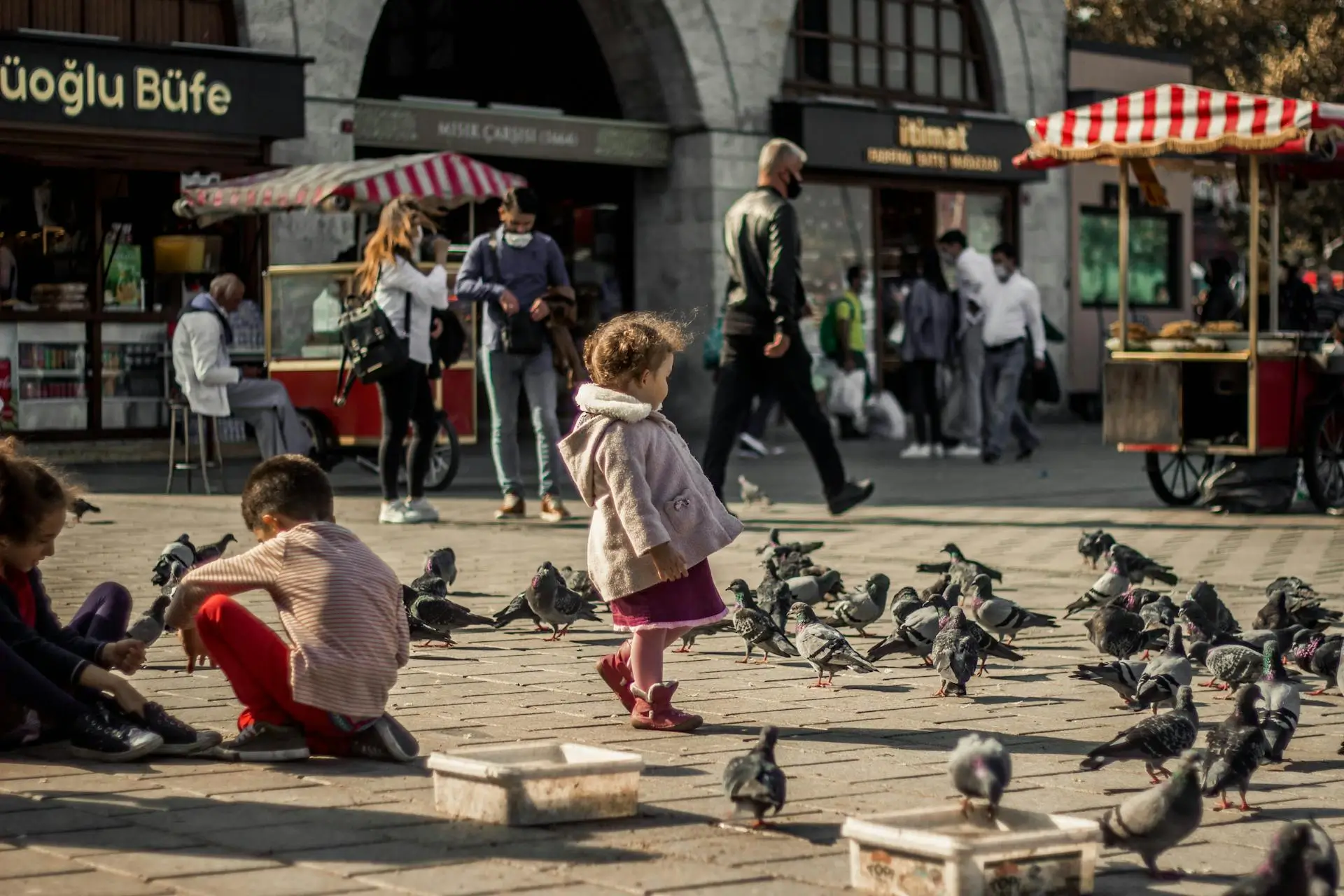 Enfant au milieu des pigeons sur une place animée d’Istanbul, avec stands de rue et passants en arrière-plan.