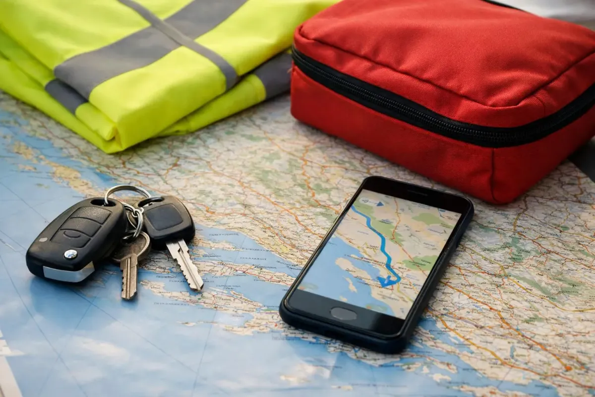 Une vue de dessus des essentiels d'un voyageur posés sur une table en bois avant un road trip en Croatie : clés de voiture, carte routière, gilet de sécurité, trousse de premiers soins et smartphone avec application de navigation, à la lumière naturelle du matin.