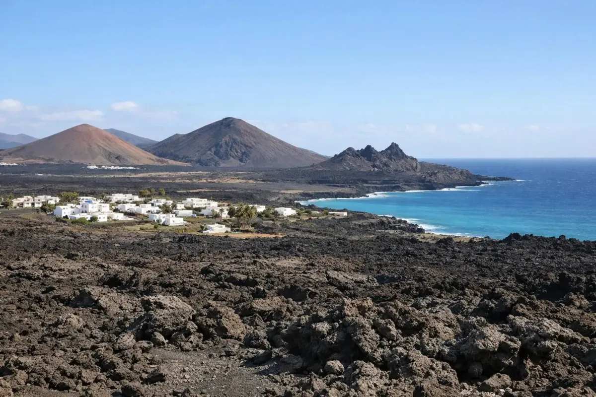 Paysage volcanique de Lanzarote montrant des champs de lave noire solidifiée, petites maisons blanches traditionnelles dispersées, océan Atlantique turquoise visible à l'horizon, formations géologiques uniques créant des textures naturelles, lumière méditerranéenne éclatante, pas de touristes, perspective grand angle capturant l'immensité du terrain volcanique