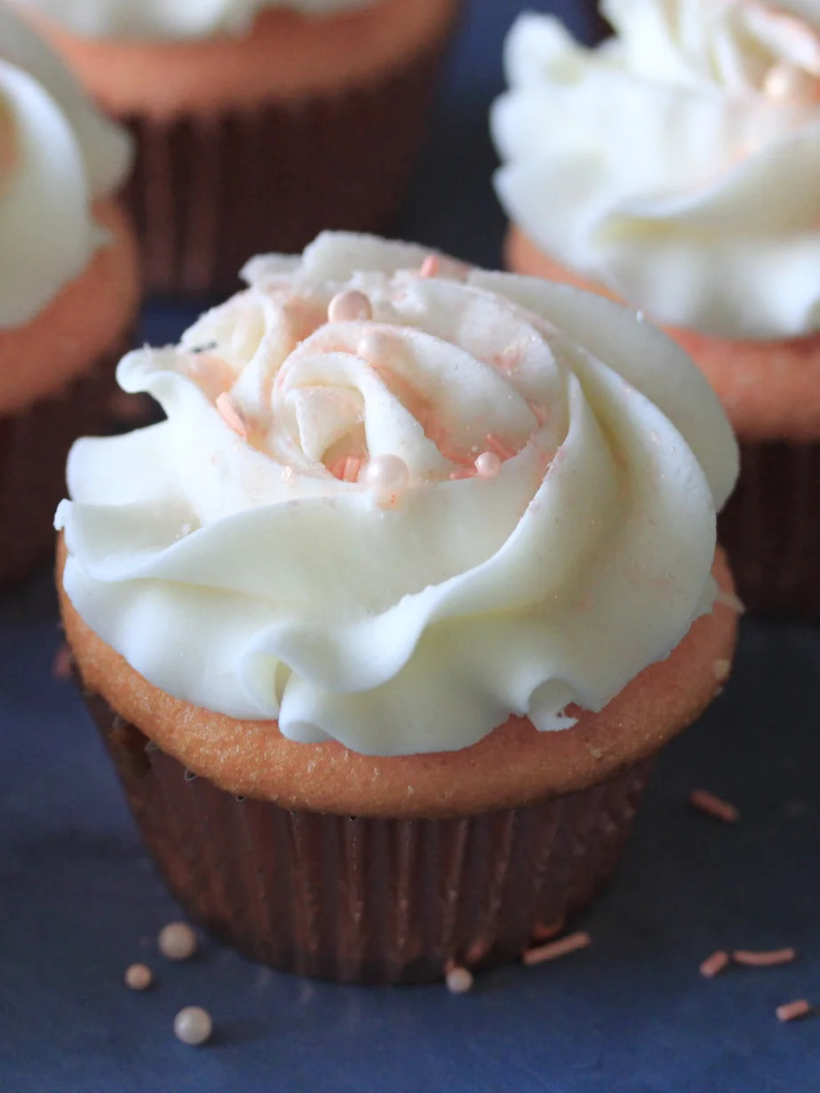 Vanilla cupcake with white swirled frosting decorated with pink sprinkles, on a dark surface with other cupcakes blurred in background.