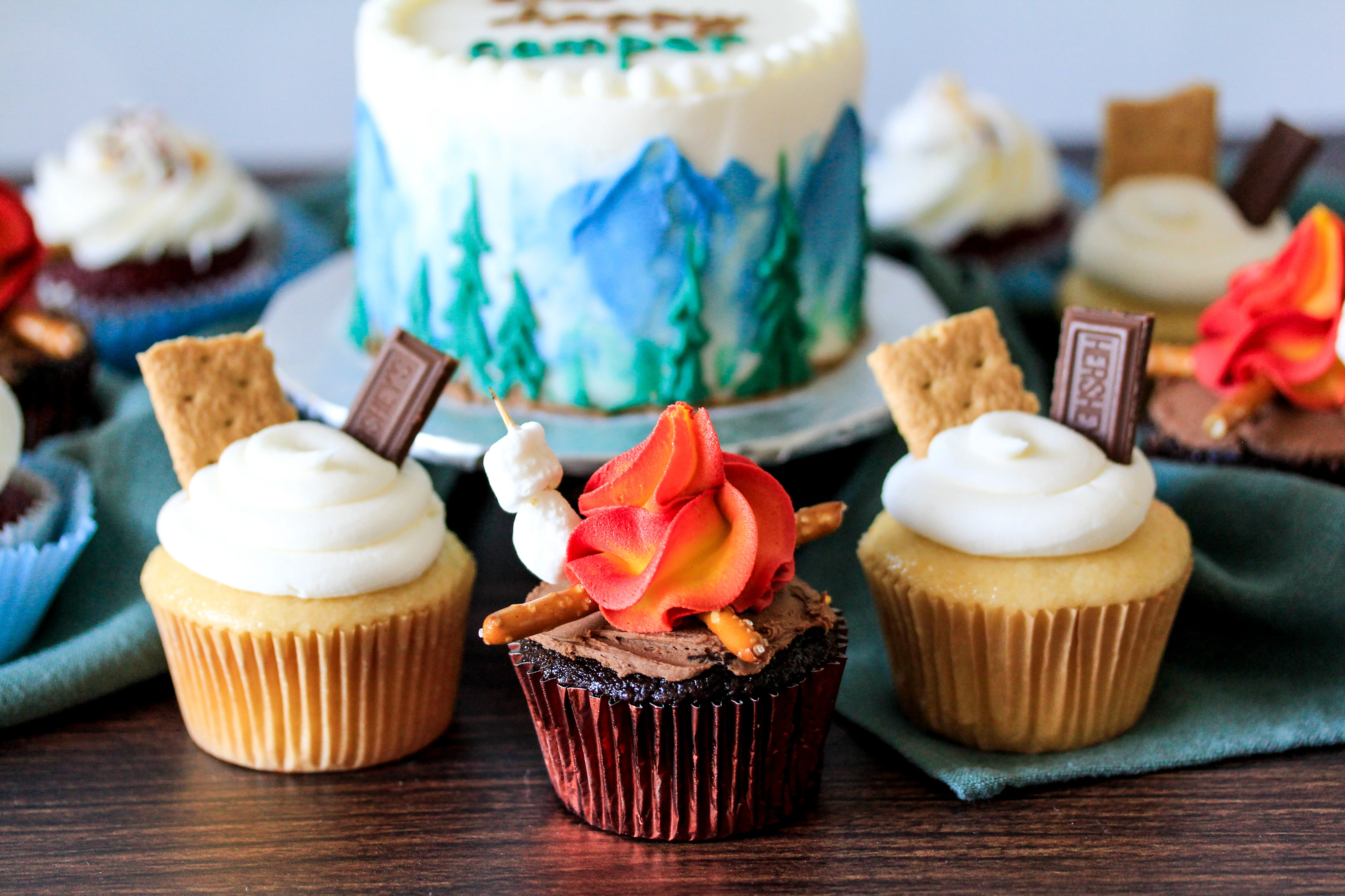 Cupcakes decorated for a campfire theme with marshmallows, pretzels, graham crackers, chocolate pieces, and flame-like red and yellow frosting, with a forest and mountain cake in the background.