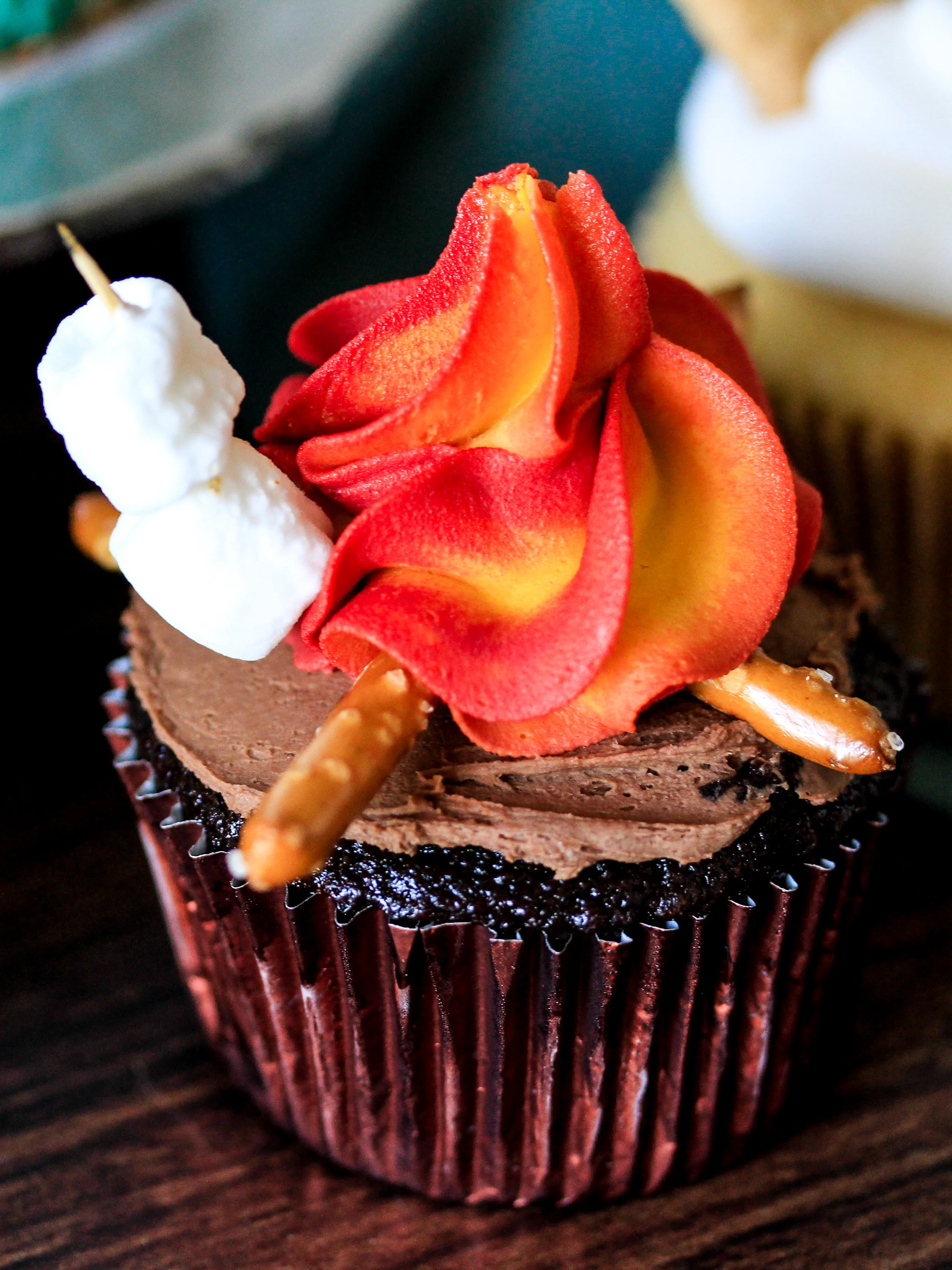 Chocolate cupcake with chocolate frosting, decorated with red and orange flame-shaped icing, pretzel sticks, and two marshmallows on a toothpick.