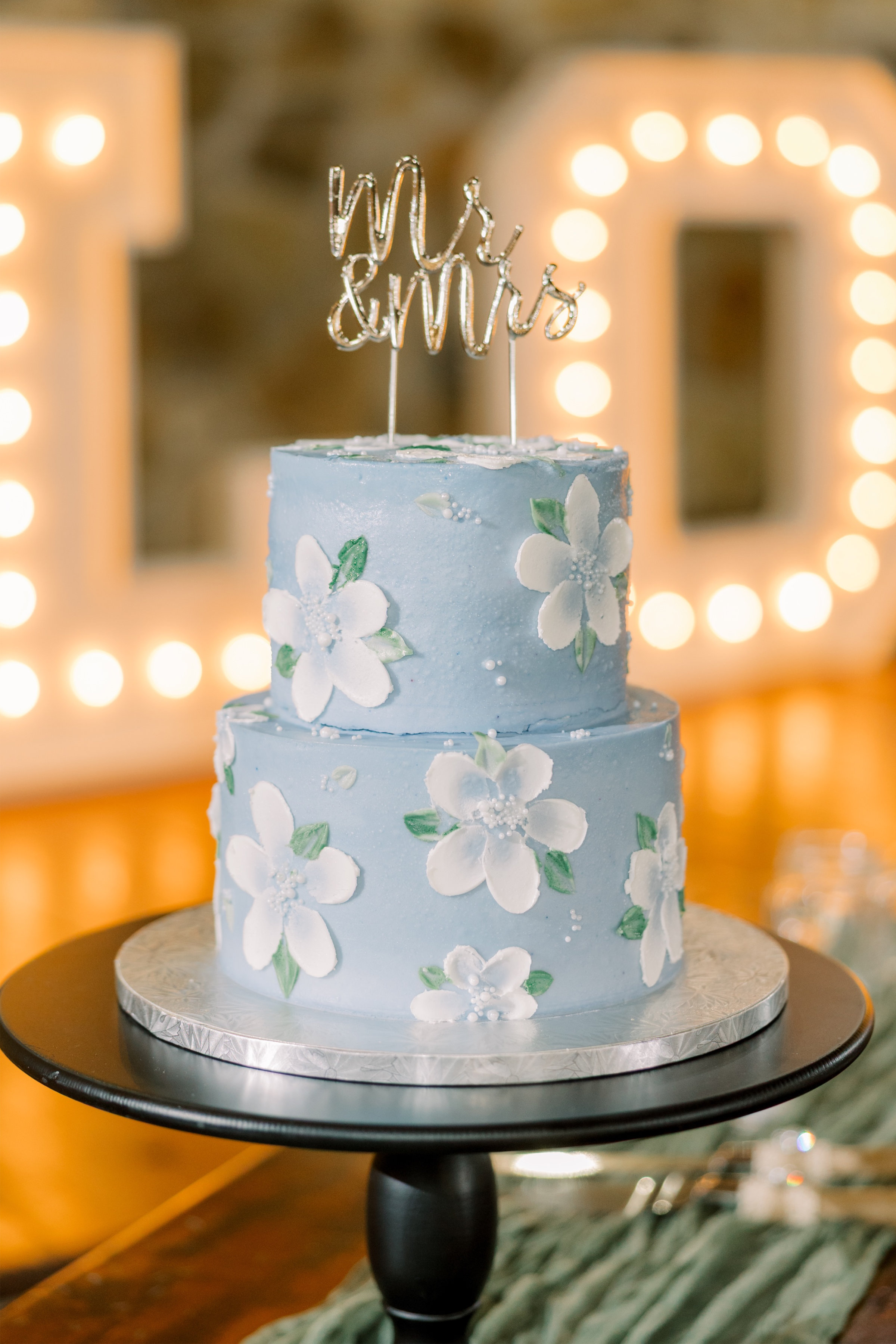 Two tier wedding cake with The Sugar Art Blueberry colored buttercream and hand painted floral accented sign at The Spings Wedding Venue in Oklahoma.