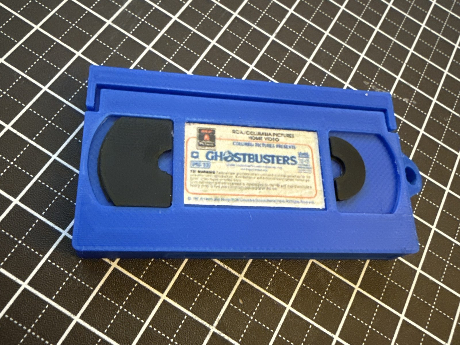 Miniature blue 3D-printed VHS tape keychain with black reels and a printed paper label, on a cutting mat