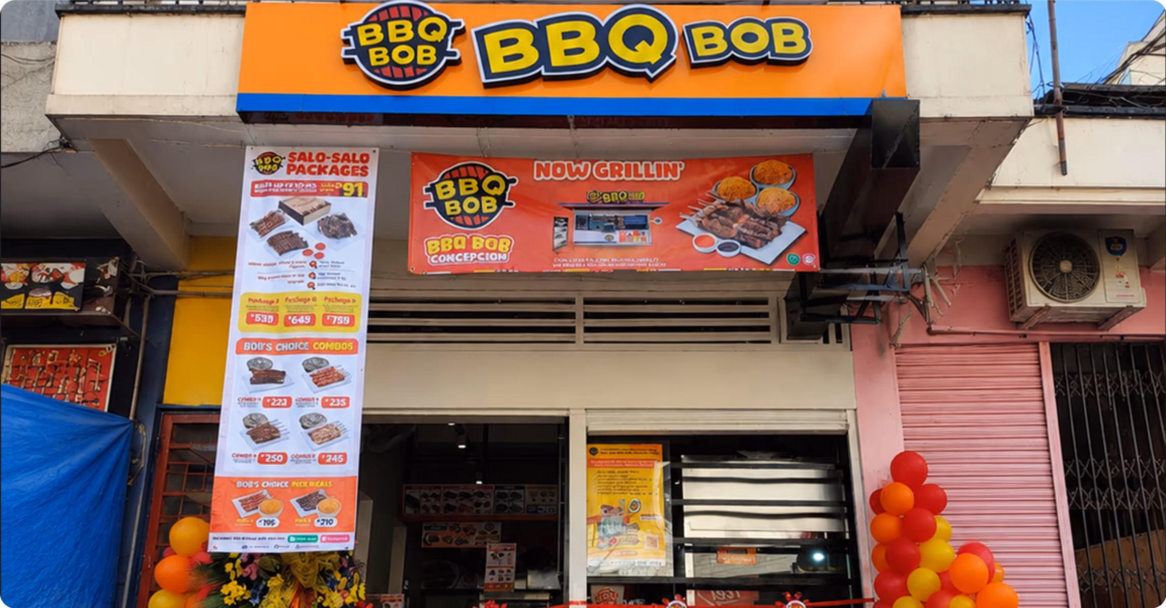 Front of BBQ Bob restaurant with orange sign, menu banners, balloons, and outdoor air conditioning unit.