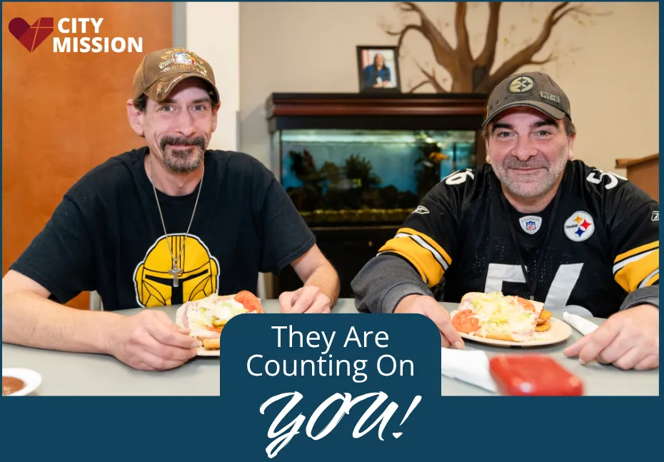 Two smiling City Mission residents in ball caps sit at a table with plates of food, beside the message They Are Counting On You.