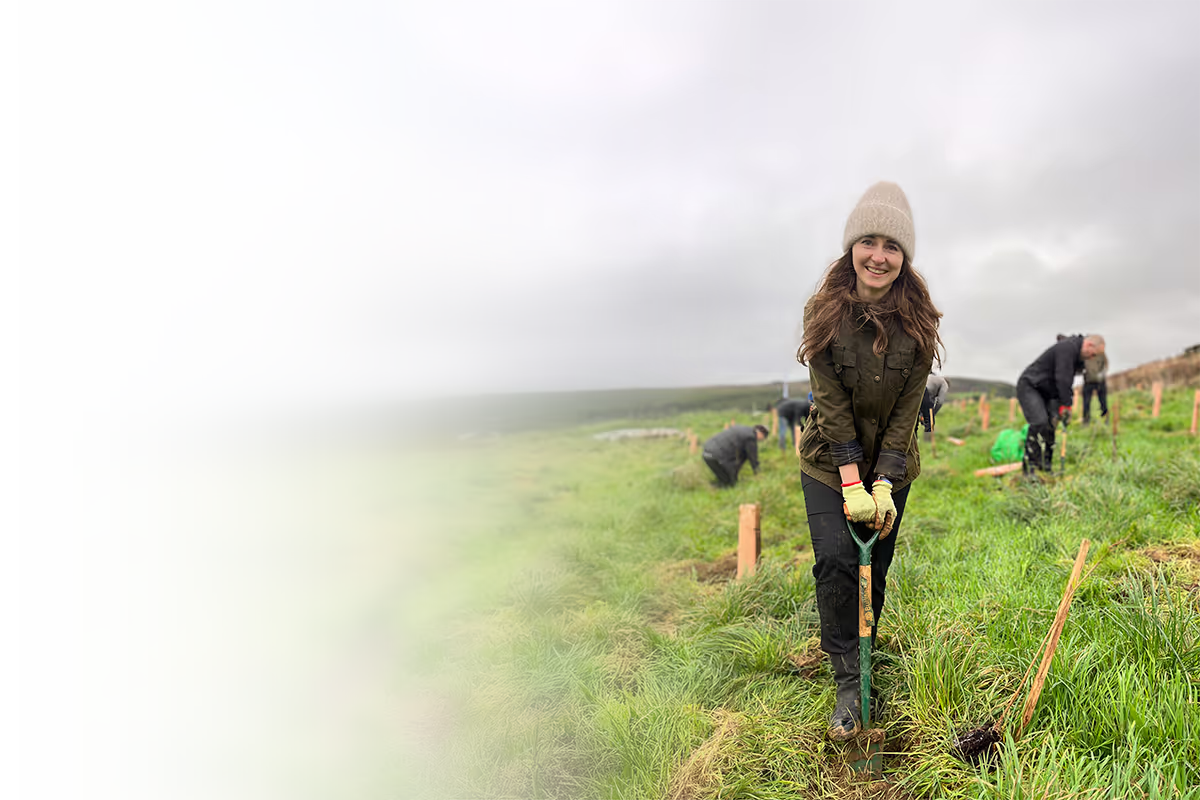 Smiling woman wearing a beanie and gloves digging with a spade in a grassy field with others planting trees under a cloudy sky.
