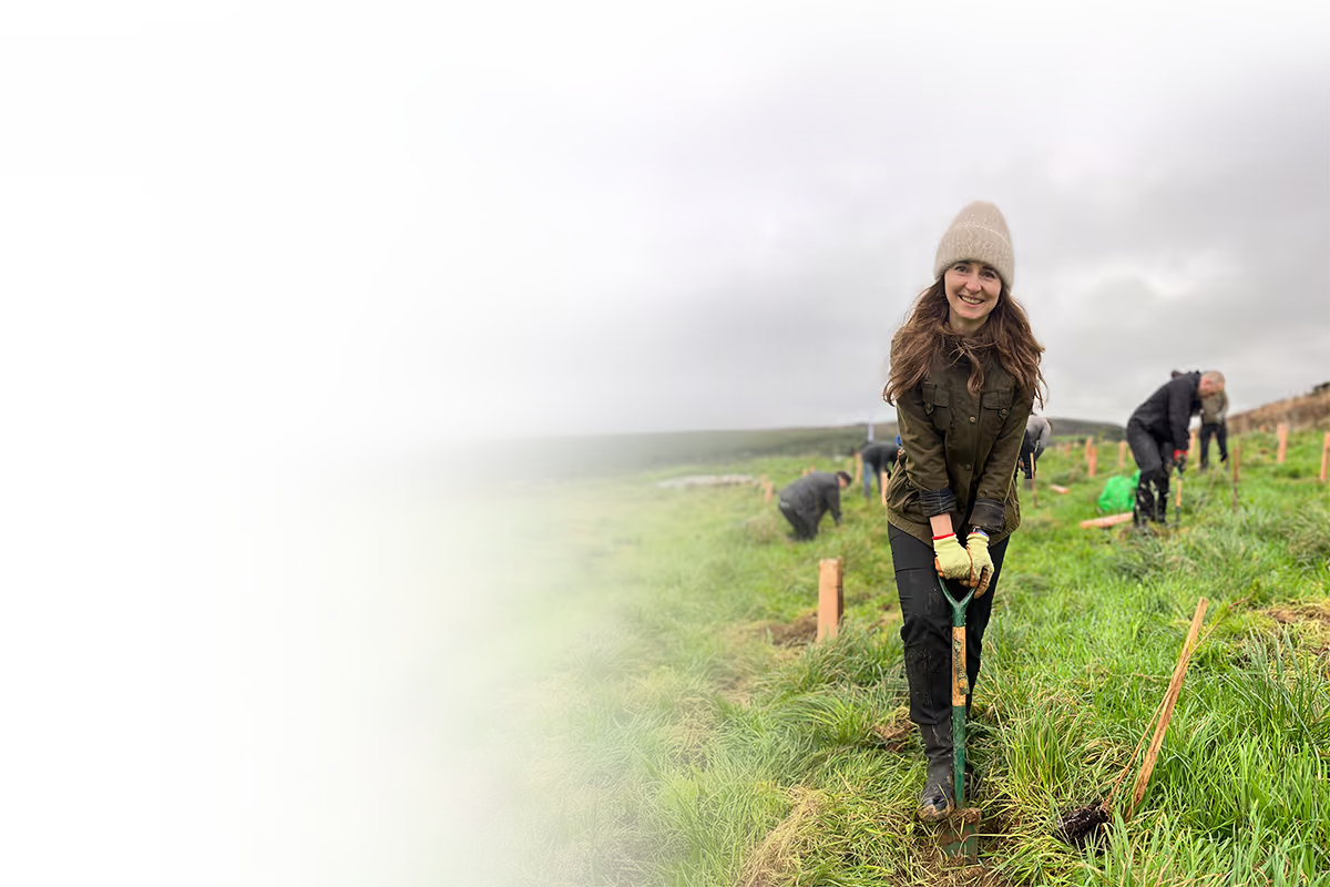 Smiling woman wearing a beanie and gloves using a shovel to plant in a grassy field with several people working in the background under a cloudy sky.
