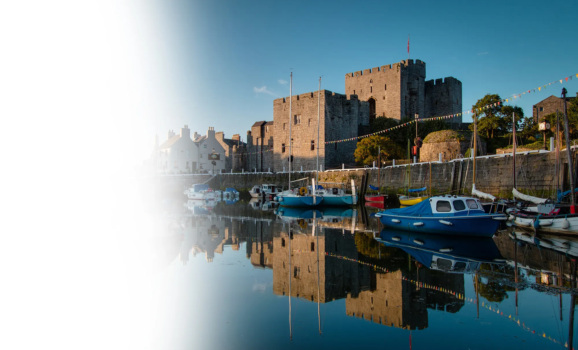 Stone castle by a calm harbor with boats reflected in the water under a clear blue sky.