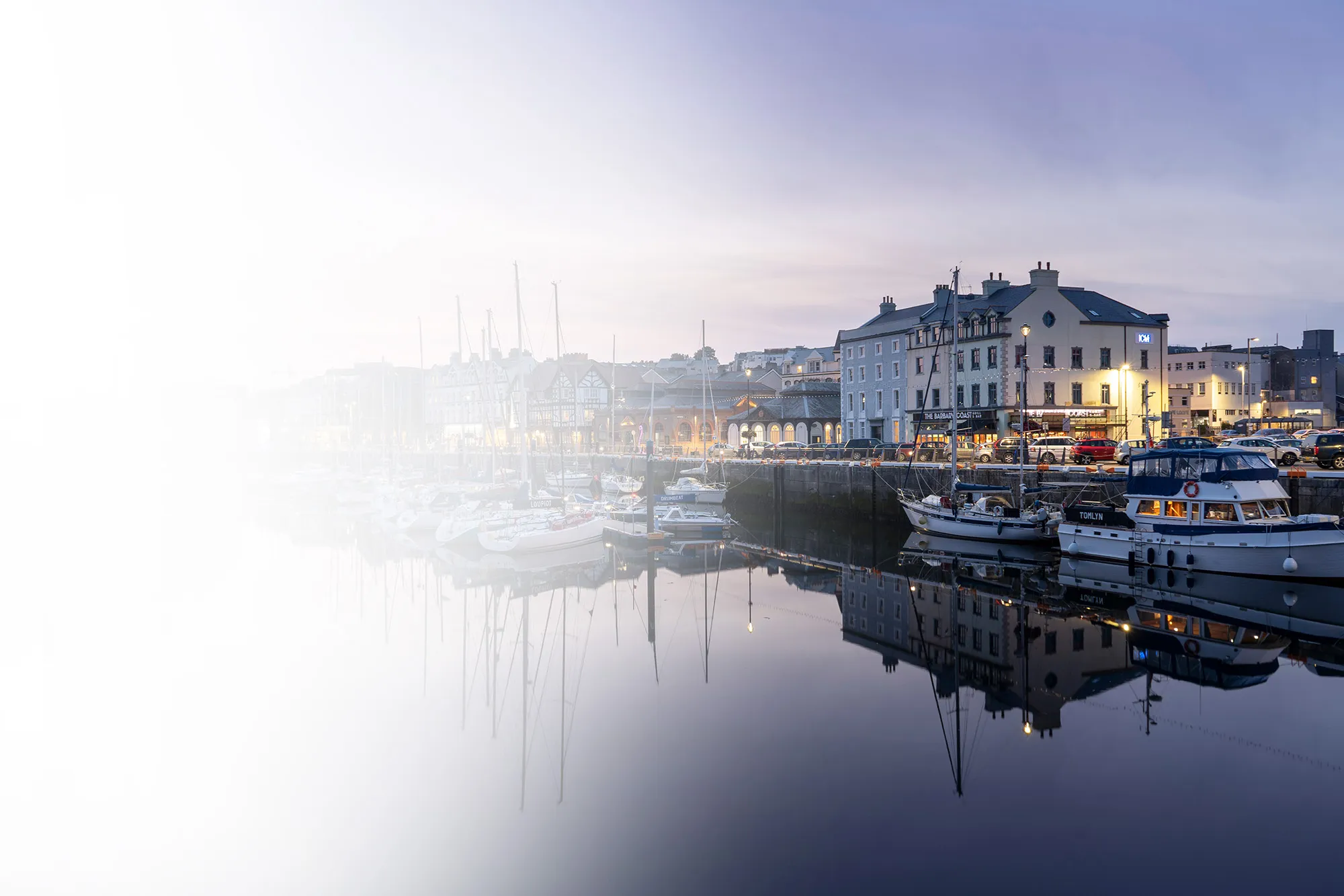 Evening view of sailboats docked at a calm harbor with illuminated buildings reflected in the water.