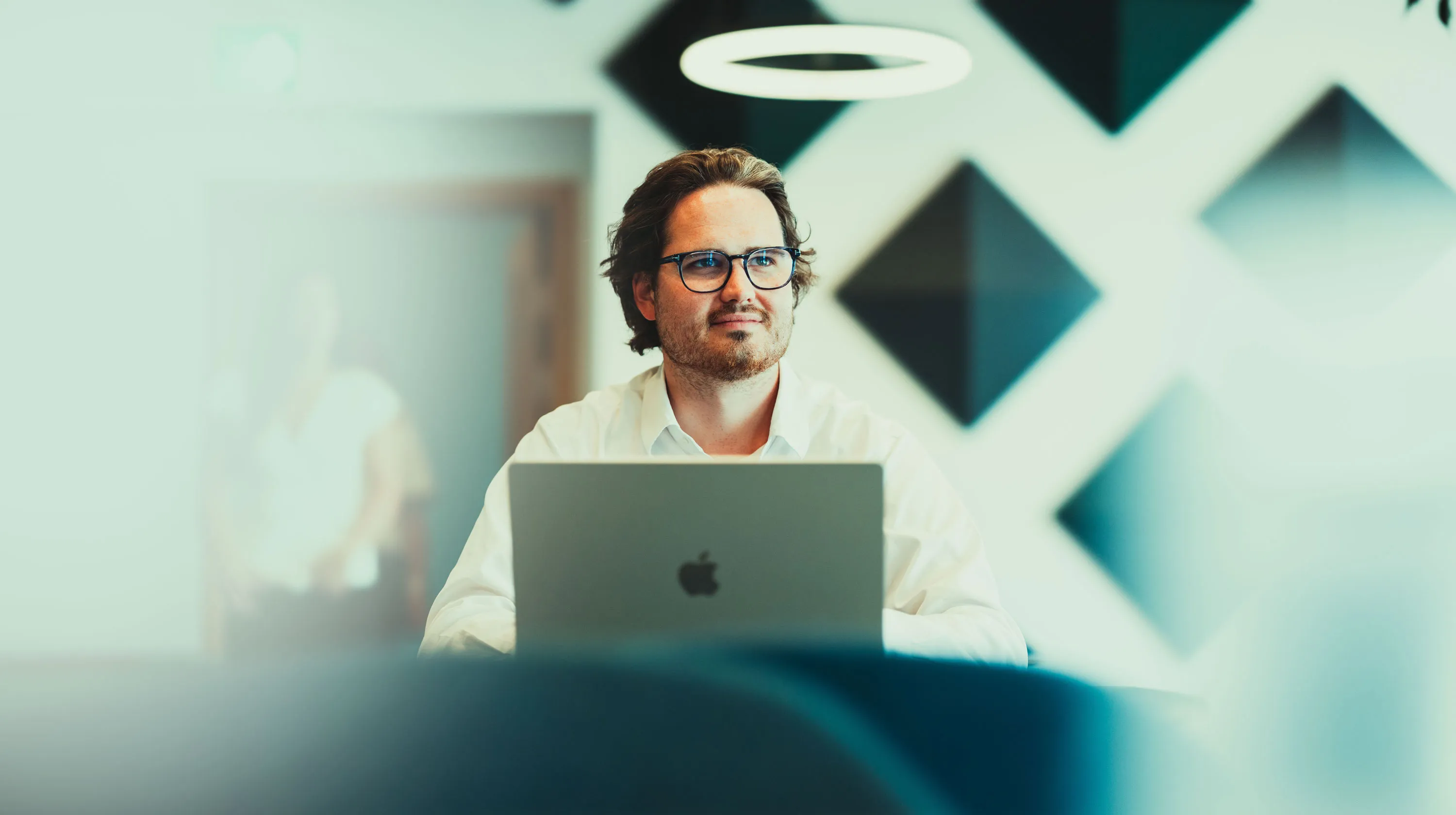 Man with glasses sitting at a desk using a MacBook laptop in a modern office space.