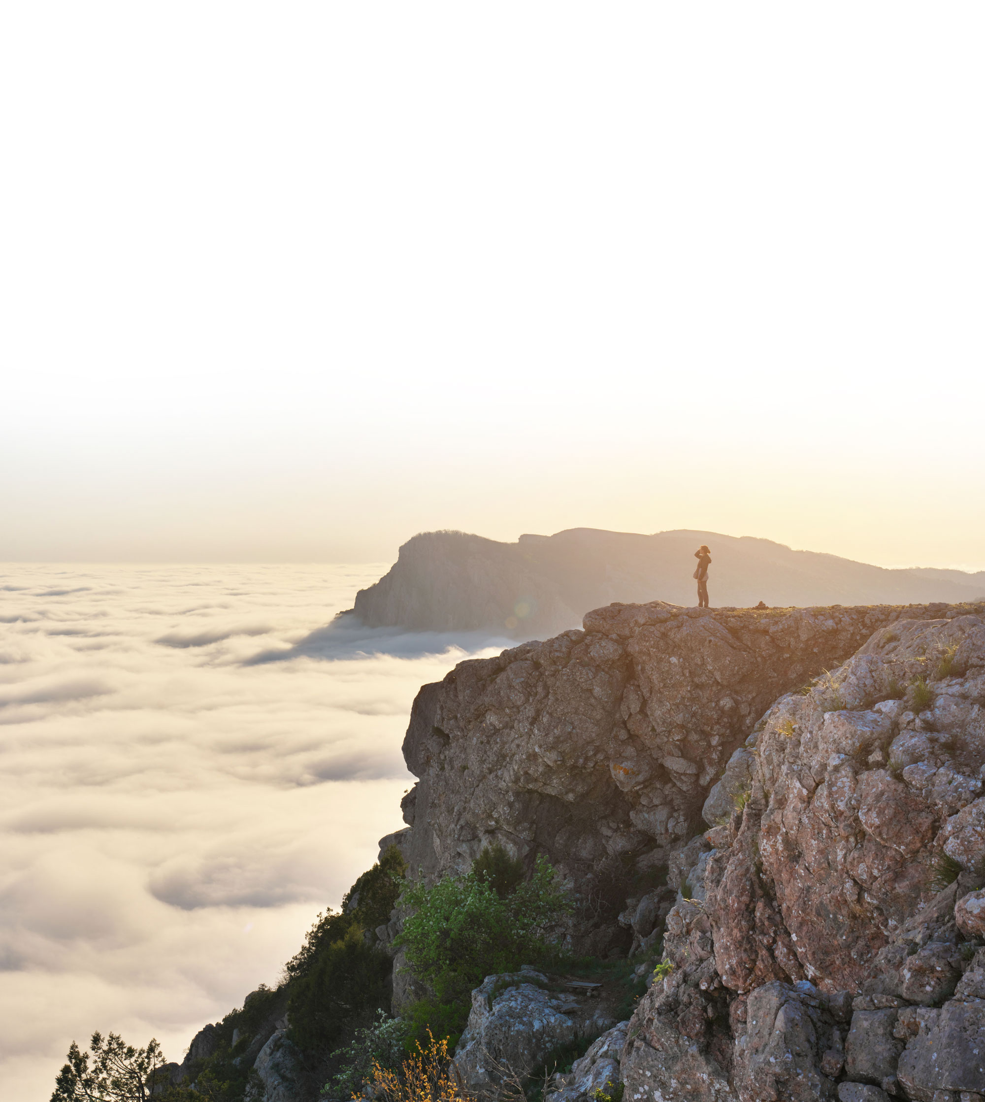 Person standing on rocky cliff edge overlooking a sea of clouds at sunrise or sunset.