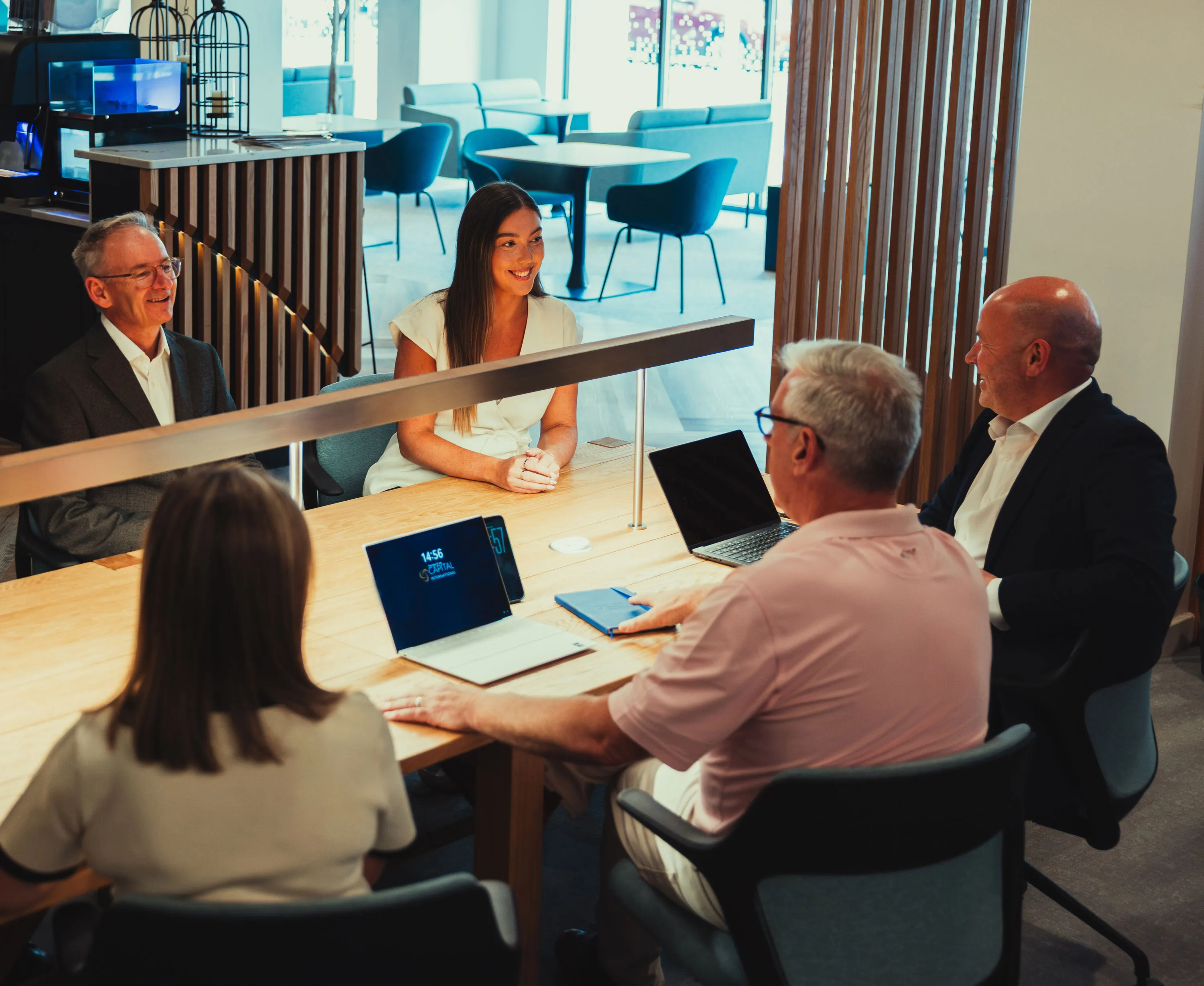 Five people sitting around a table in a modern office having a meeting with laptops and notebooks.