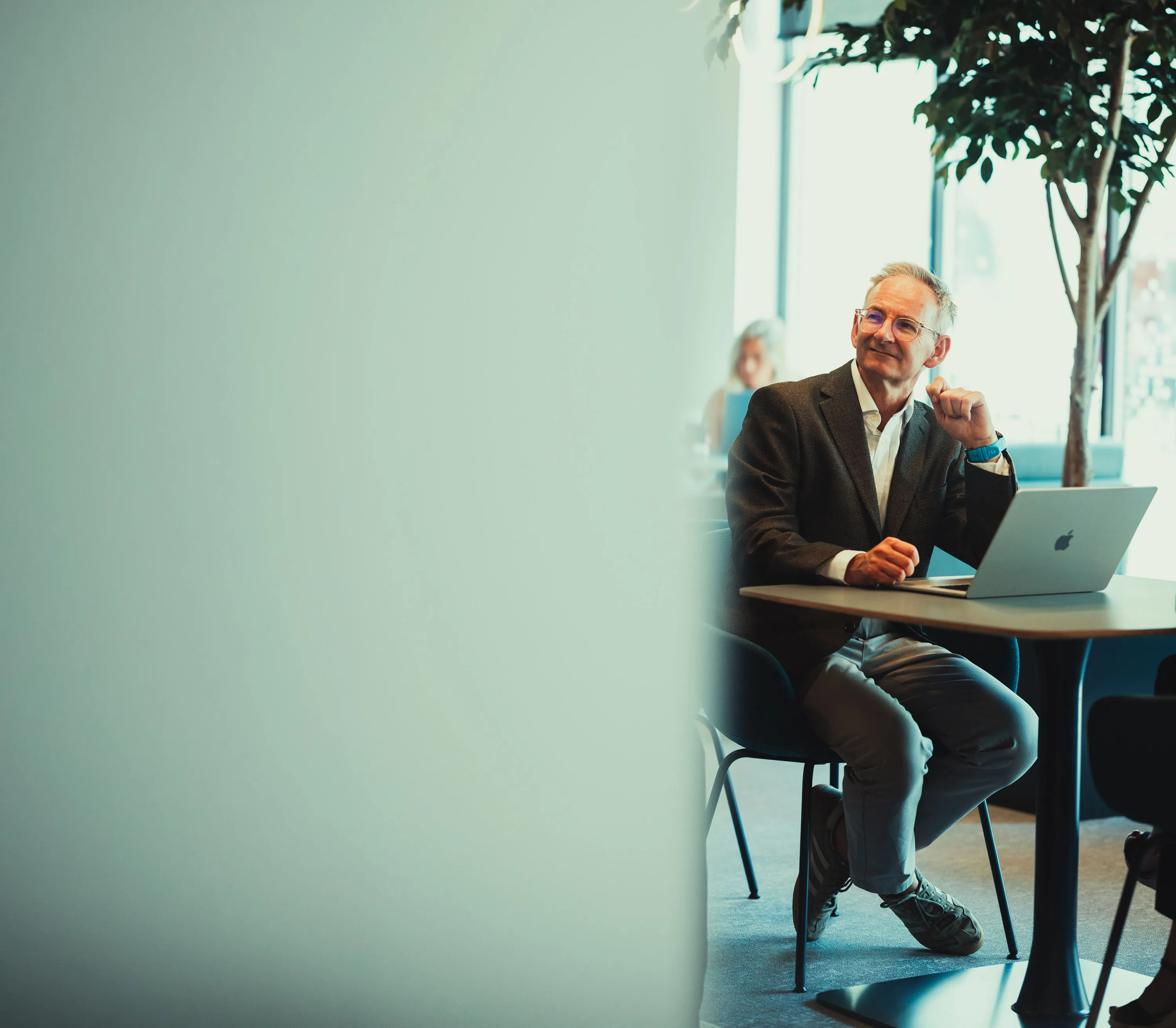 Man in glasses and blazer sitting at table with laptop in modern office with indoor plants.