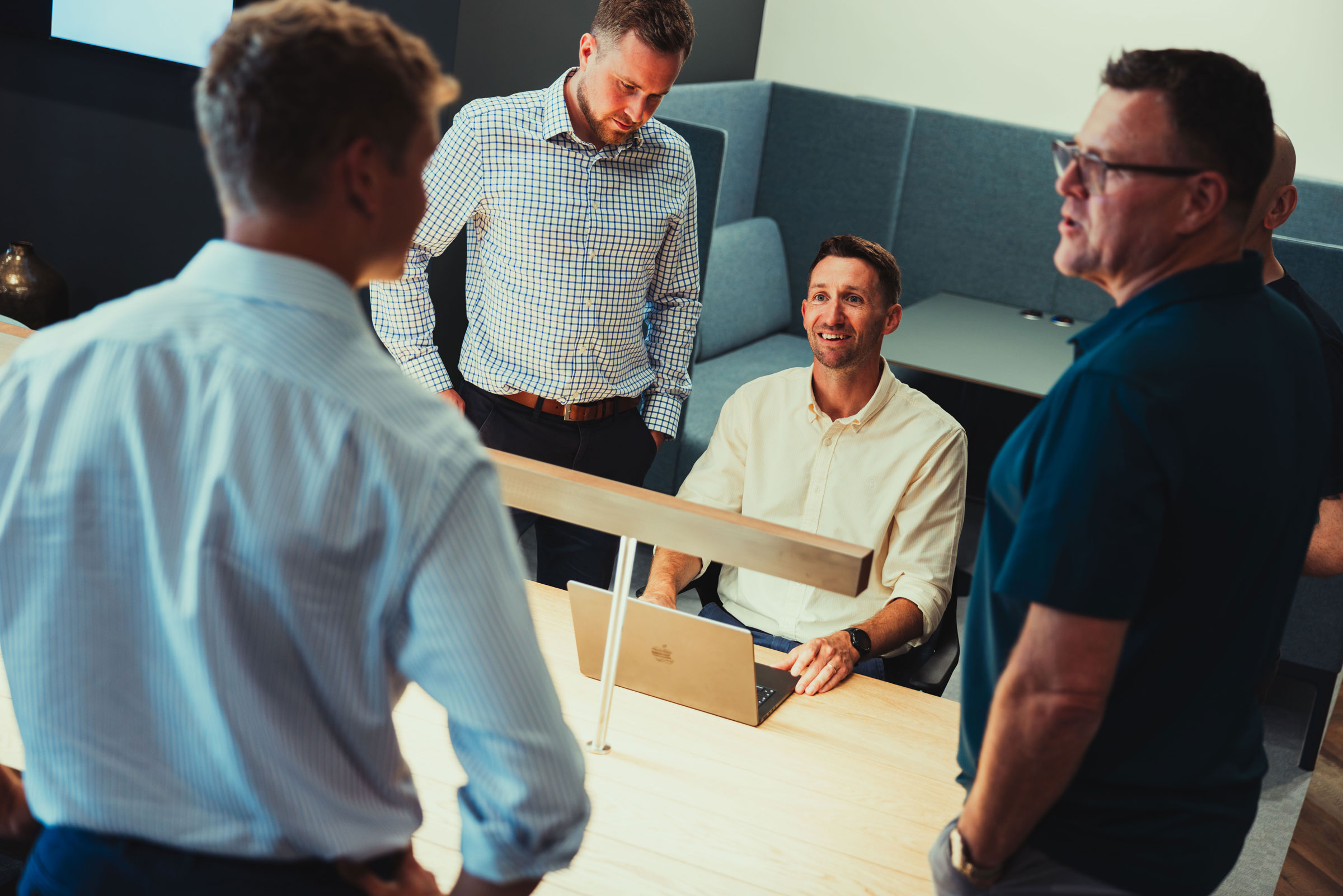 Five men in a modern office gather around a table with a laptop, engaged in discussion.