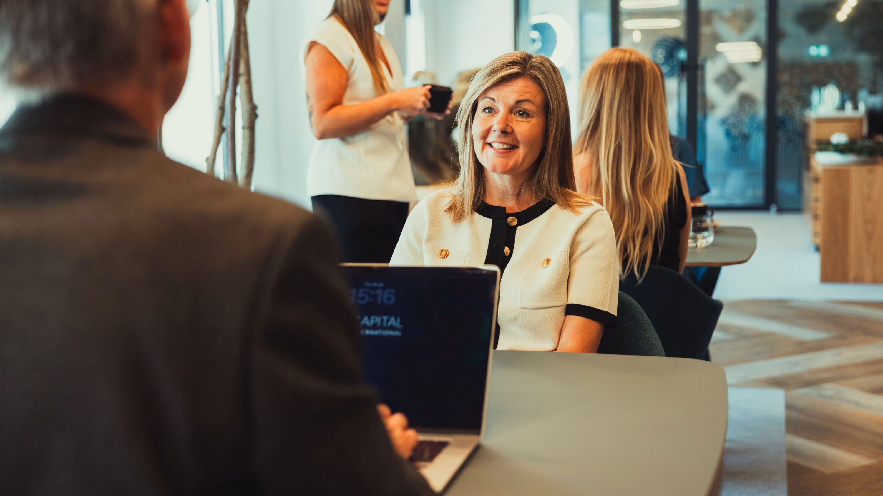 Smiling woman speaking to a man with a laptop in a modern office setting with people in background.
