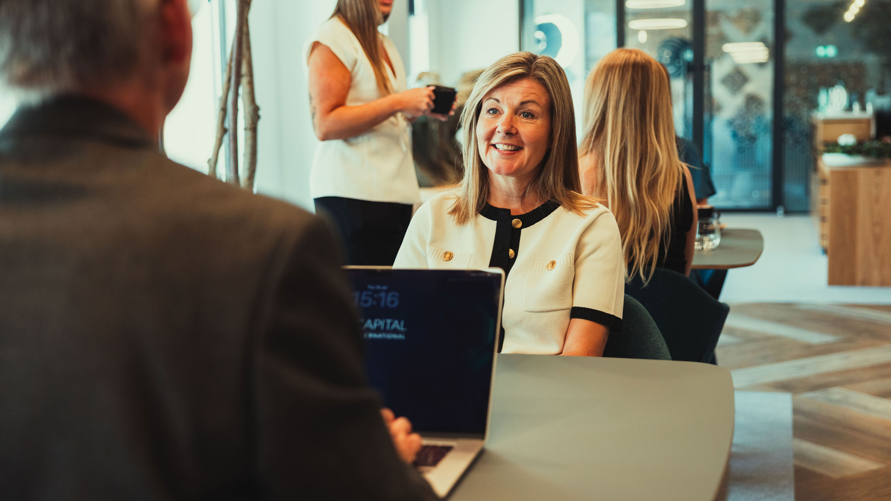 Smiling woman speaking to a man with a laptop in a modern office setting with people in background.