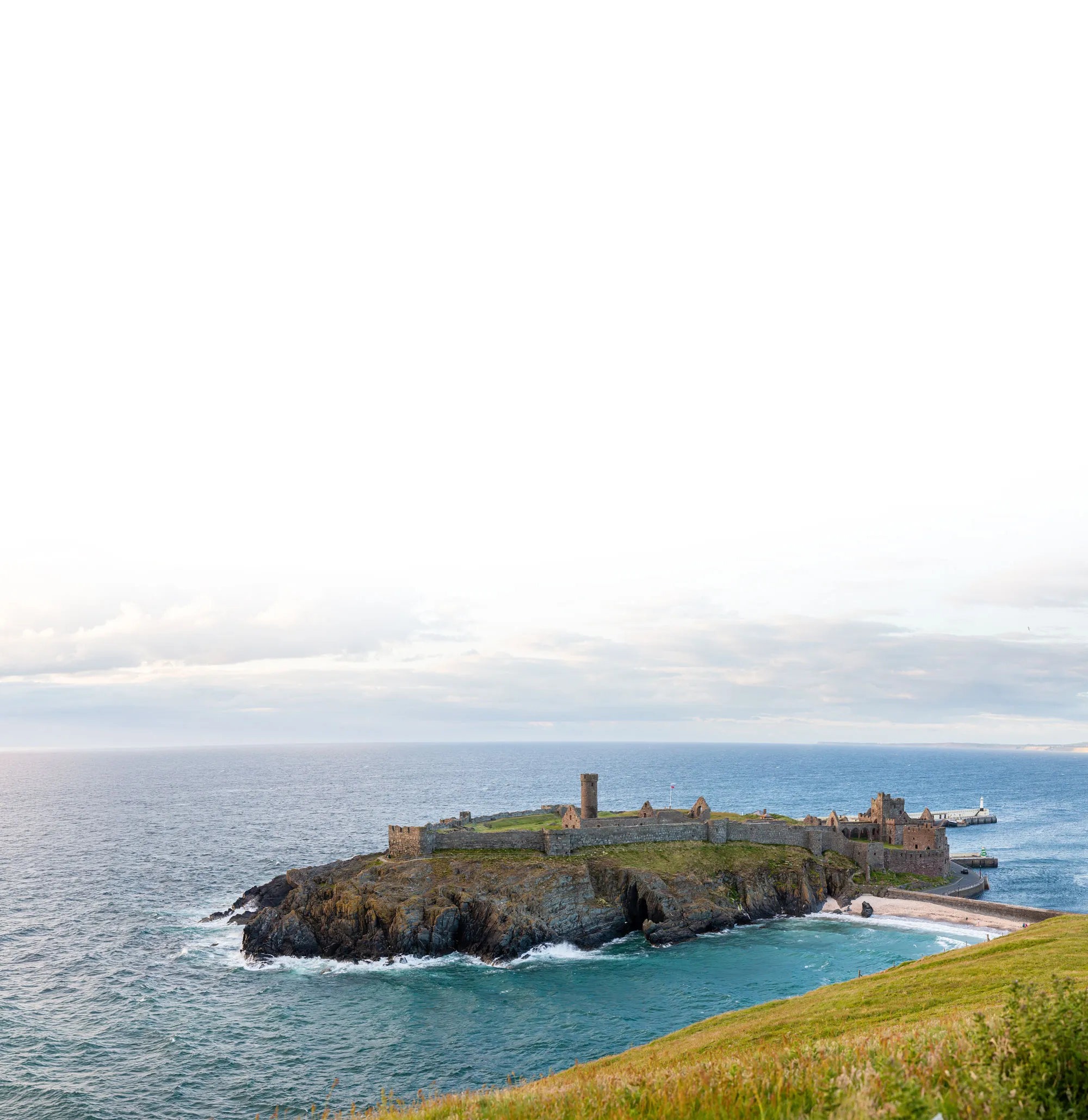 Old stone castle ruins on rocky peninsula with surrounding blue ocean and grassy foreground.