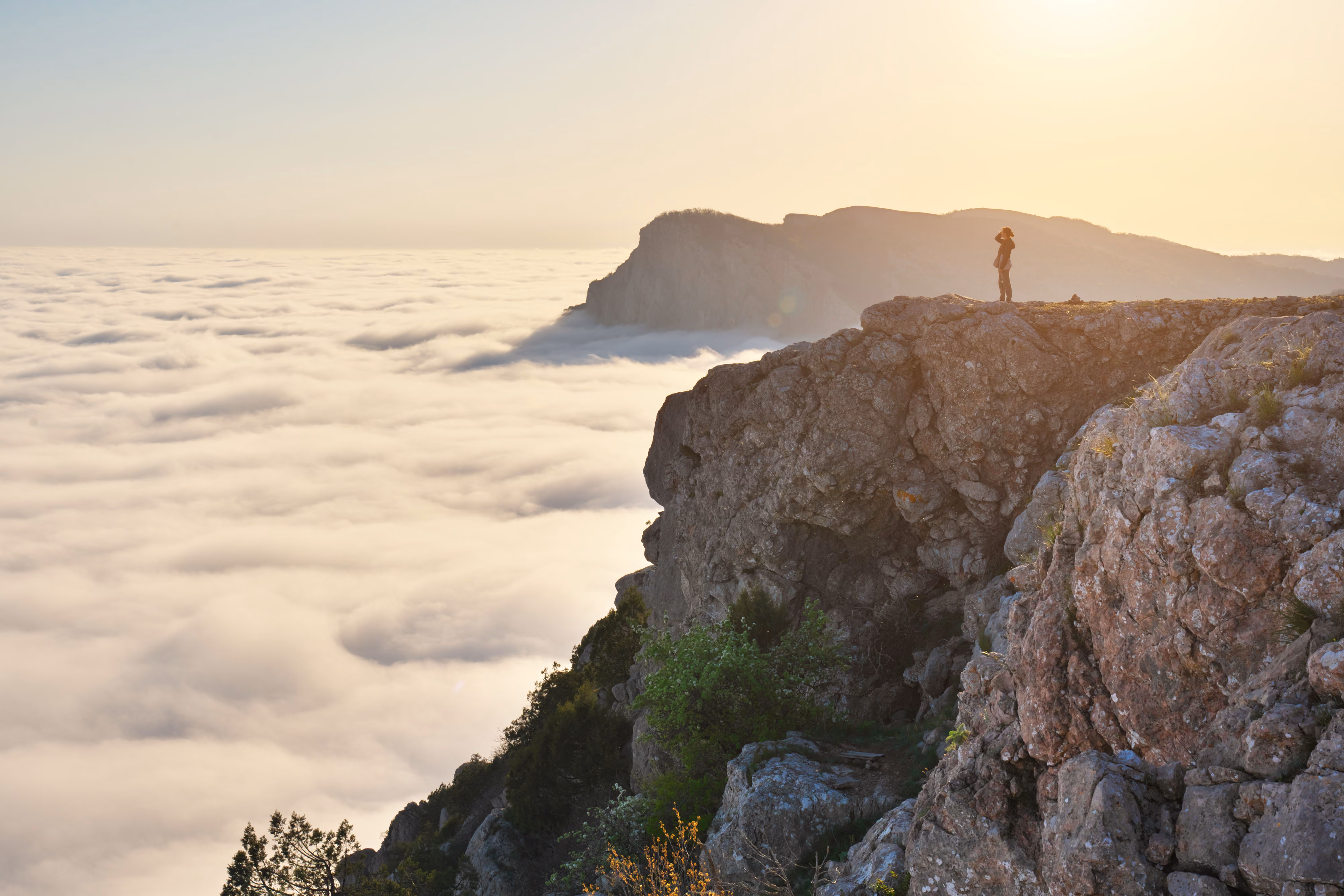 Person standing on rocky cliff overlooking a sea of clouds with distant mountains at sunrise.