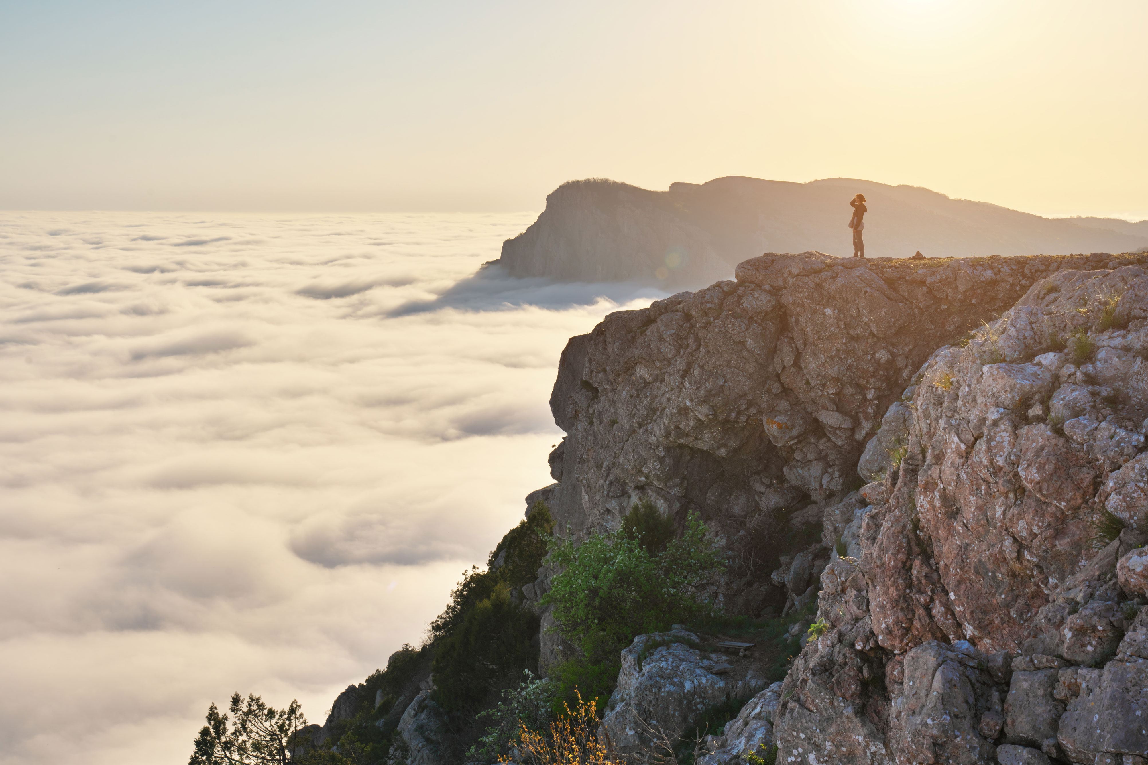 Person standing on rocky cliff overlooking a sea of clouds with distant mountains at sunrise.