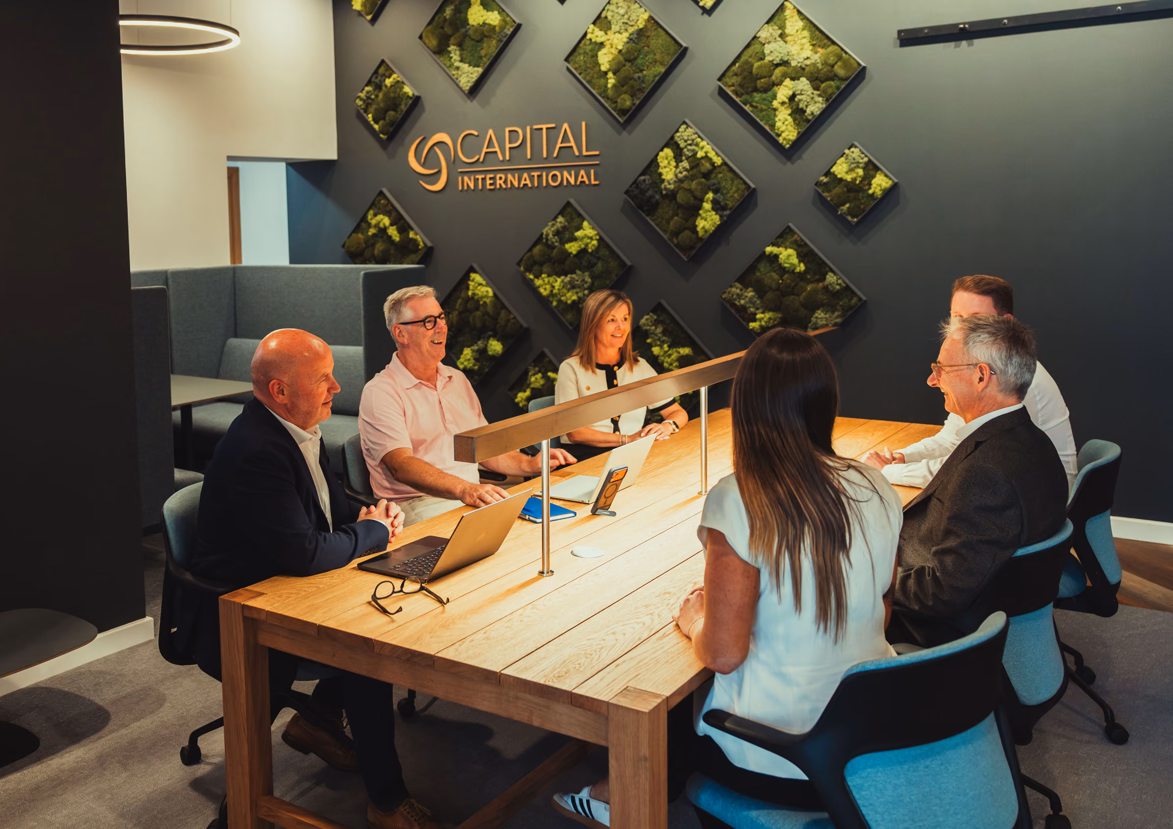 Six people seated around a wooden conference table in an office with green wall decor and Capital International logo.