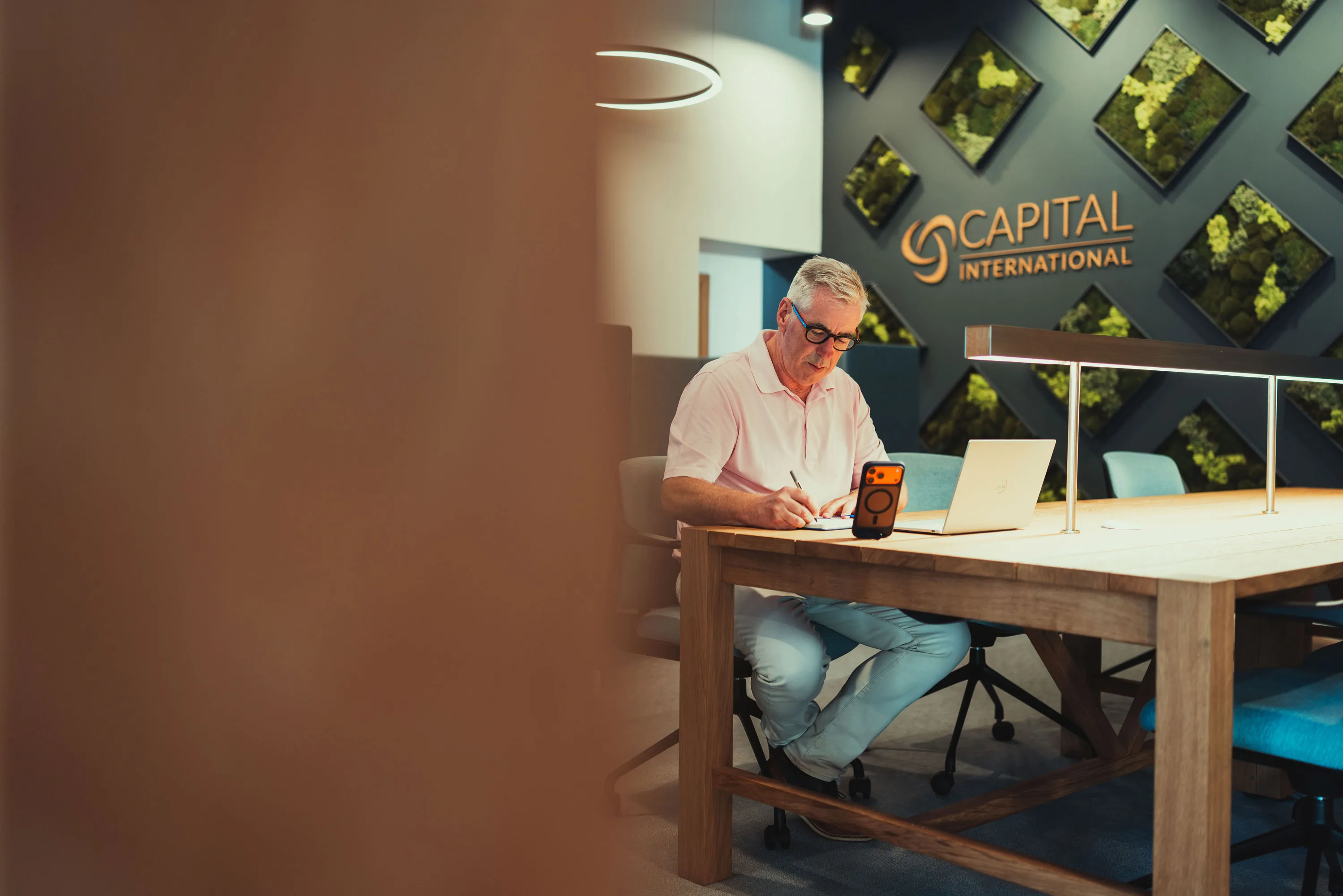 Man in light shirt writing at a wooden table with laptop and phone in a modern office space.