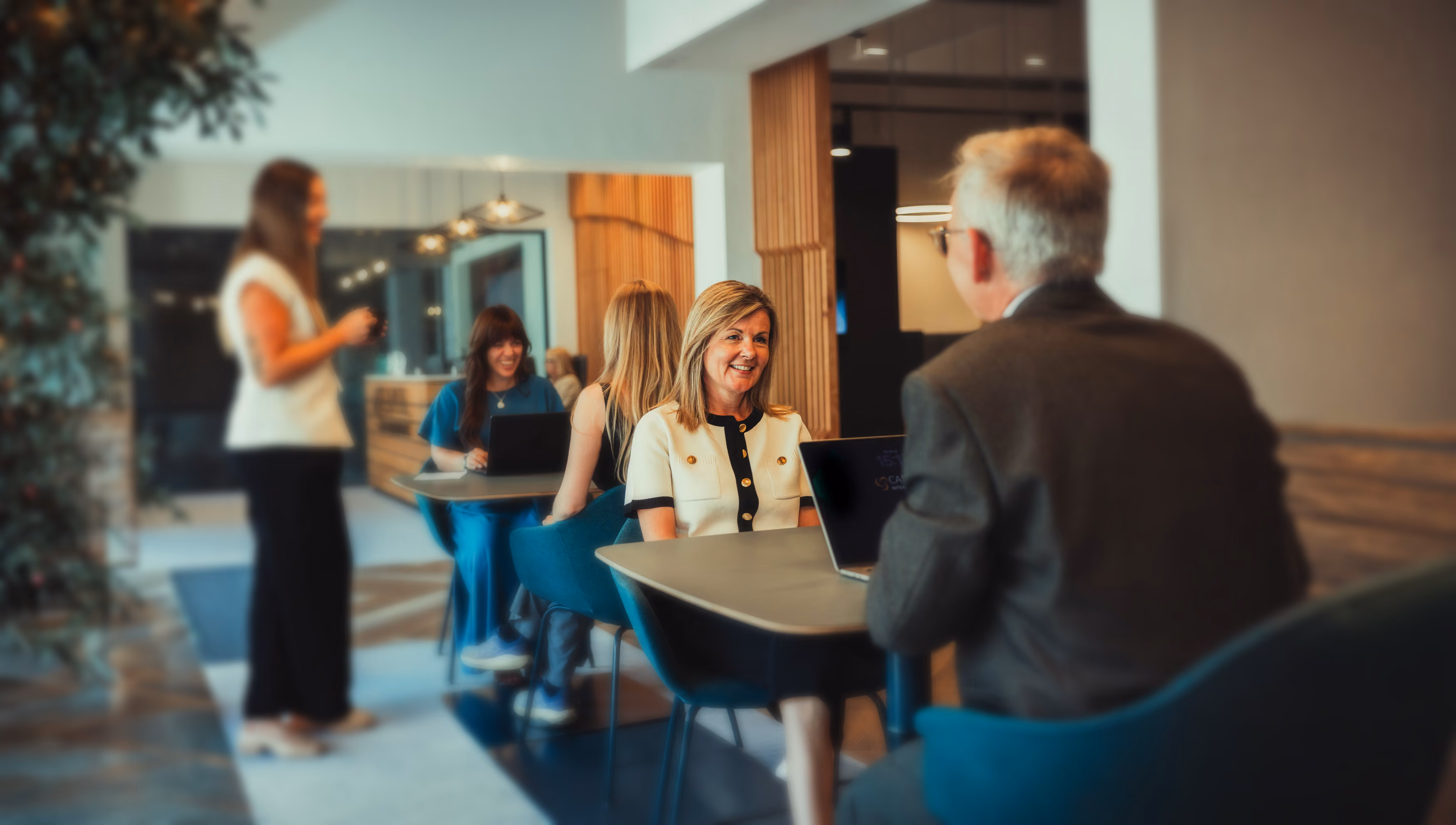 Smiling woman in white top talks with man across table in modern co-working space with others nearby.