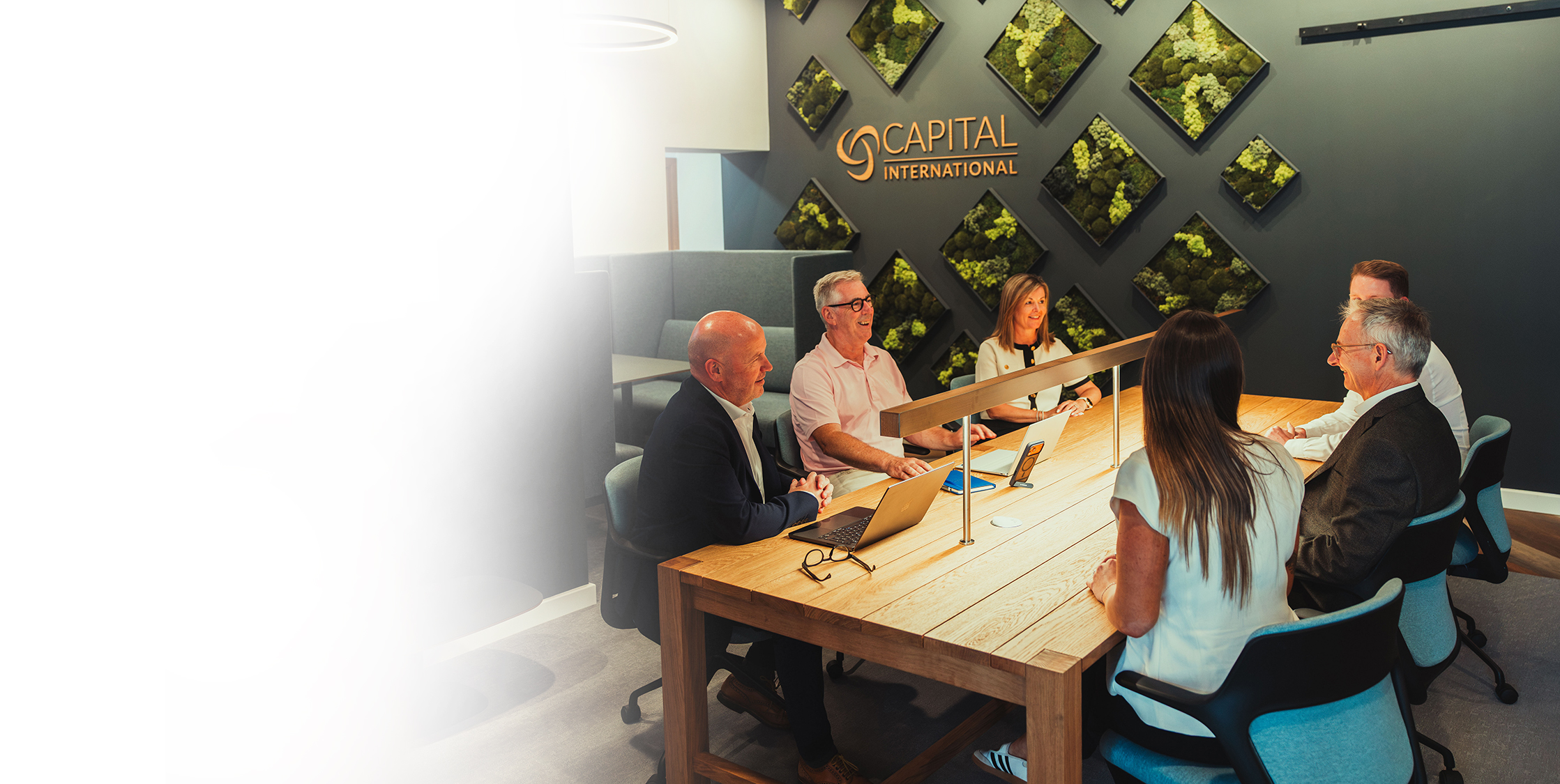 Six people having a meeting around a wooden table in a modern office with green wall art.