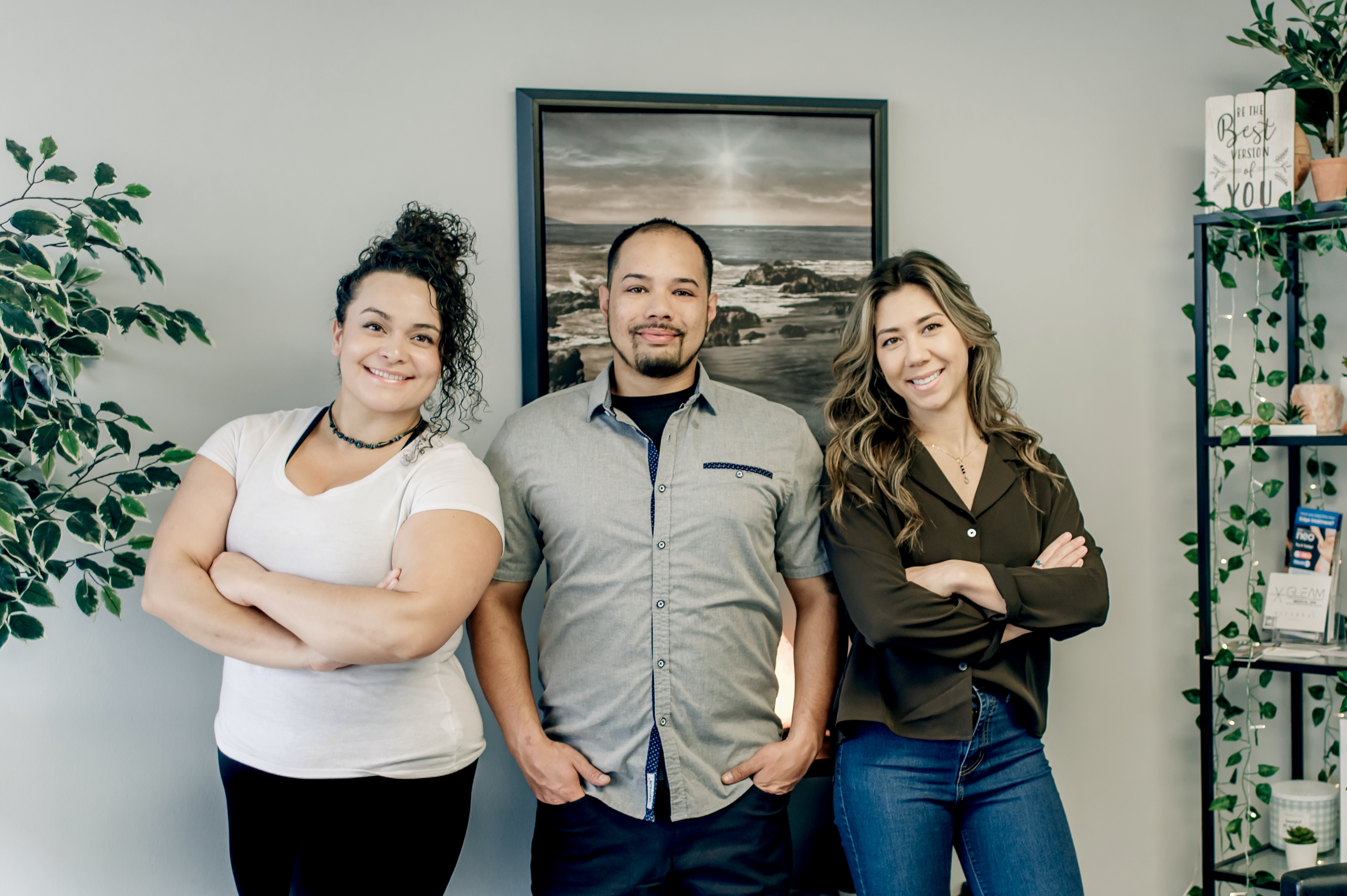 Three smiling people standing indoors with arms crossed, posing in front of a framed ocean painting and plants.